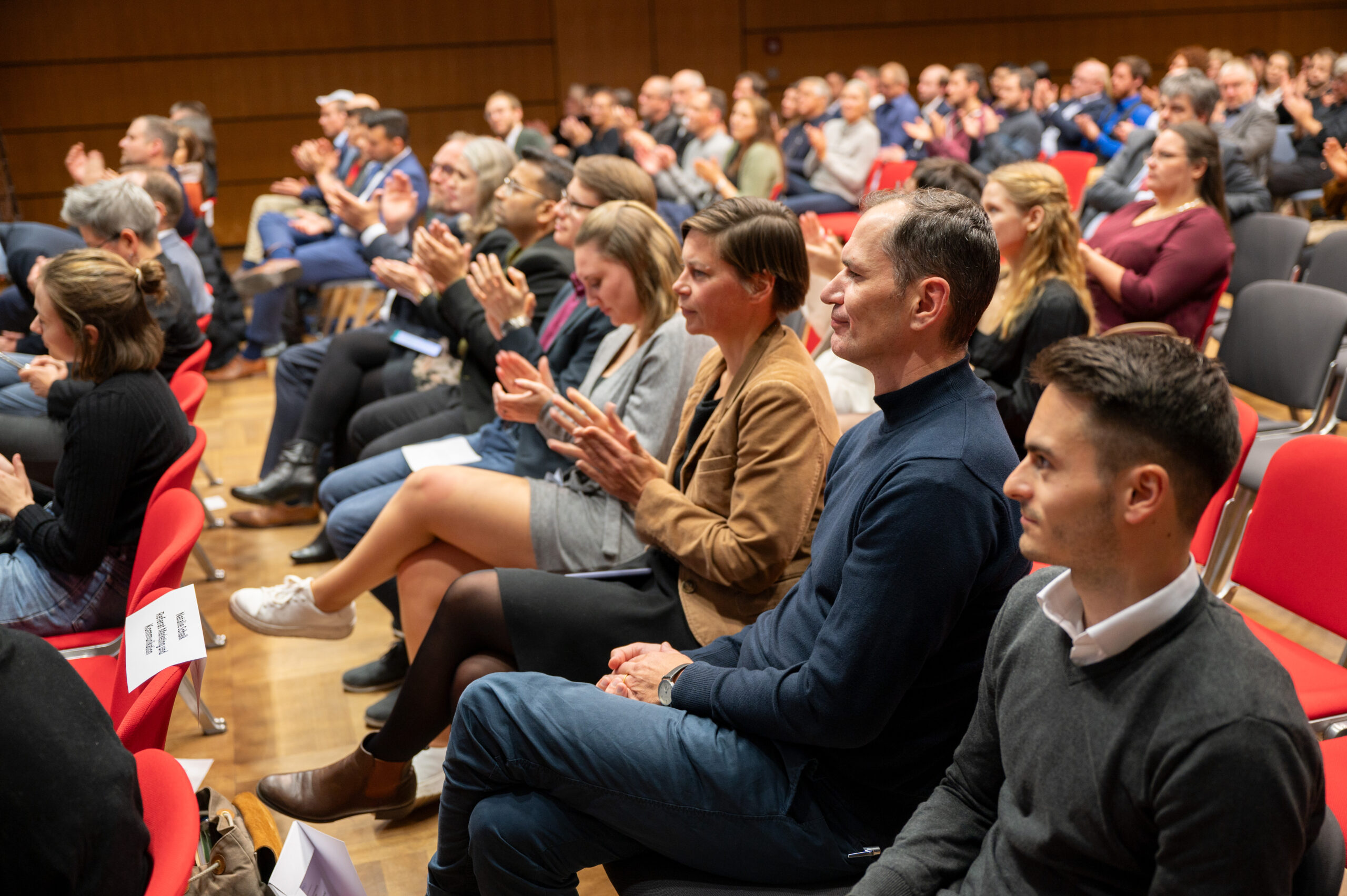 Eine bunt gemischte Gruppe von Menschen sitzt in Reihen in einem Hörsaal der Hochschule Coburg, hört aufmerksam zu und klatscht. Manche tragen Freizeitkleidung, andere Business-Kleidung. Die Atmosphäre lässt auf eine formelle Veranstaltung oder Präsentation schließen.