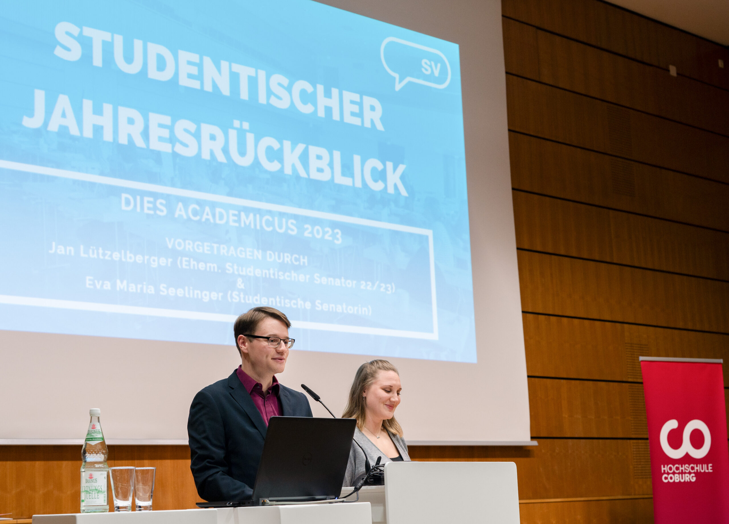 Zwei Vortragende stehen während der Veranstaltung „Studentischer Jahresrückblick“ der Hochschule Coburg an einem Podium. Auf einem Bildschirm werden Einzelheiten zur akademischen Versammlung 2023 angezeigt. Neben dem Podium ist auf einem roten Banner das Logo der Universität zu sehen, das ihr stolzes Erbe betont.