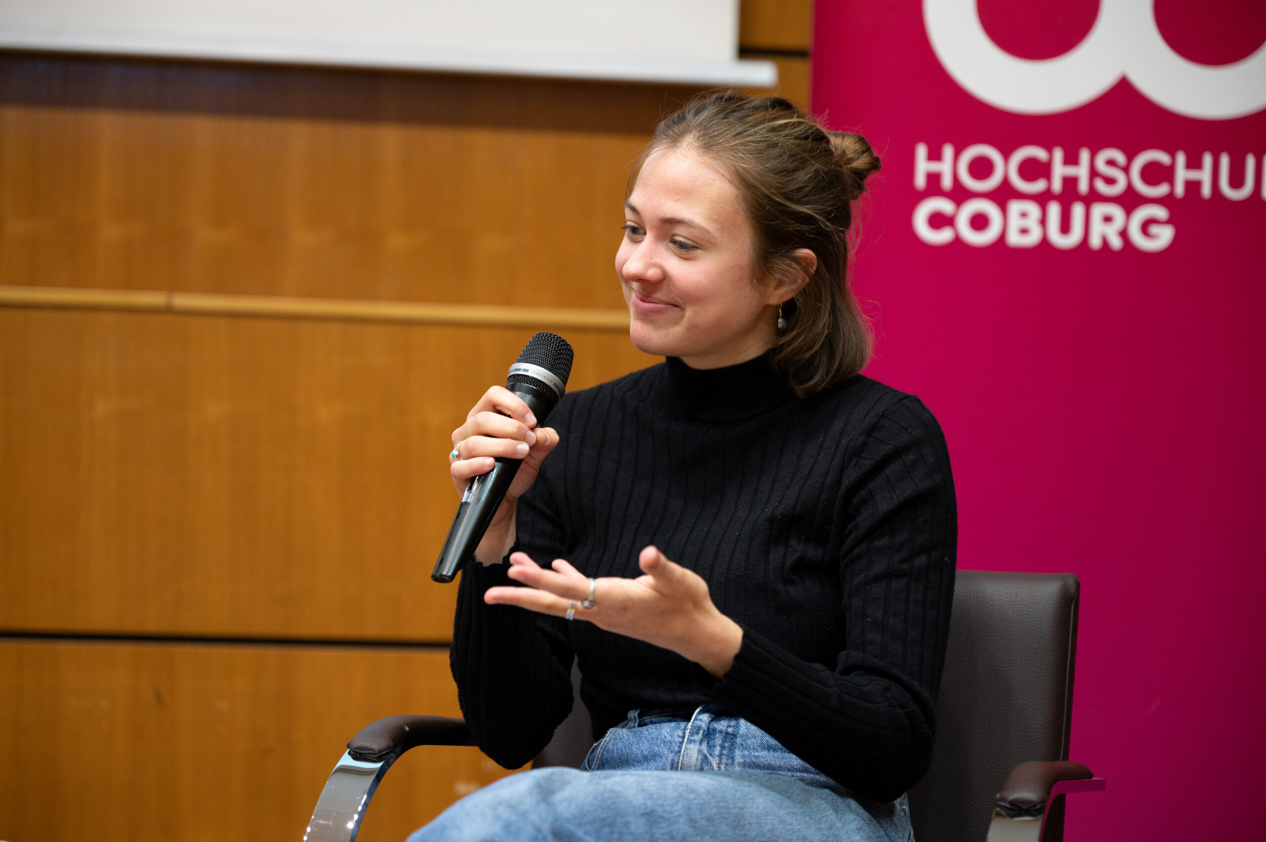 Eine Frau in schwarzem Rollkragenpullover und Jeans hält ein Mikrofon, spricht angeregt und gestikuliert mit der anderen Hand. Sie sitzt vor einem rosa Hintergrund und erzählt dem Publikum voller Leidenschaft von der Hochschule Coburg.