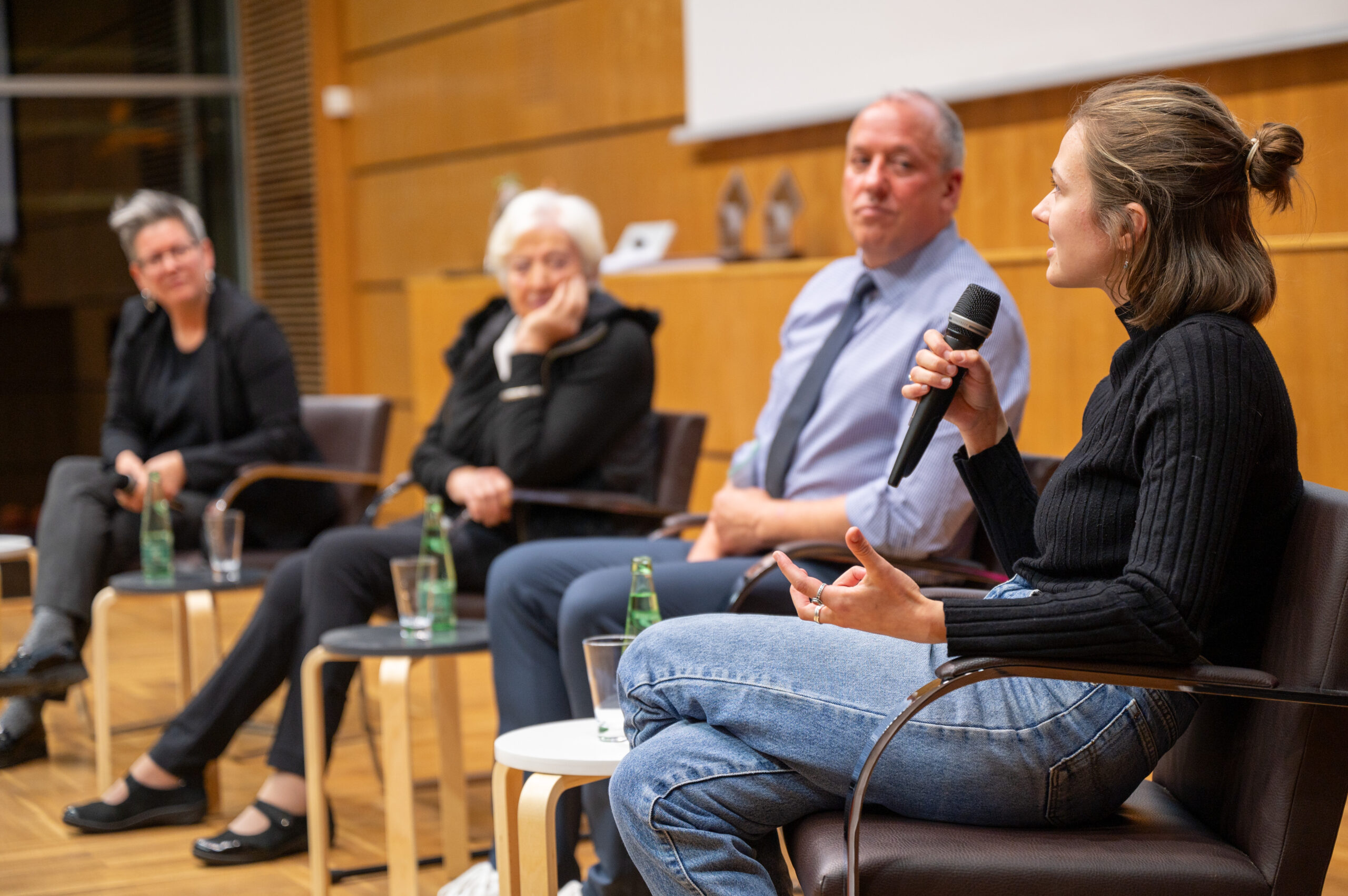 Eine Frau mit Mikrofon spricht während einer Podiumsdiskussion an der Hochschule Coburg. Sie sitzt vor drei weiteren Personen – zwei Frauen und einem Mann – in einem Raum, der wie ein Hörsaal oder Konferenzraum mit Holzvertäfelung aussieht.