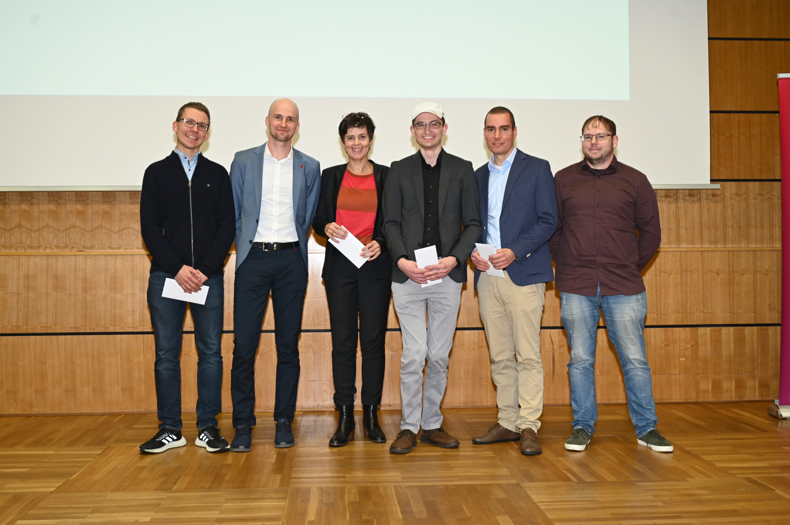 Sechs Personen stehen gemeinsam in einem Innenbereich auf einem Holzboden vor einer Holzwand. Sie sind in legerer Business-Kleidung gekleidet und halten weiße Papiere in den Händen. Sie lächeln in die Kamera und verkörpern damit den kollaborativen Geist der Hochschule Coburg.