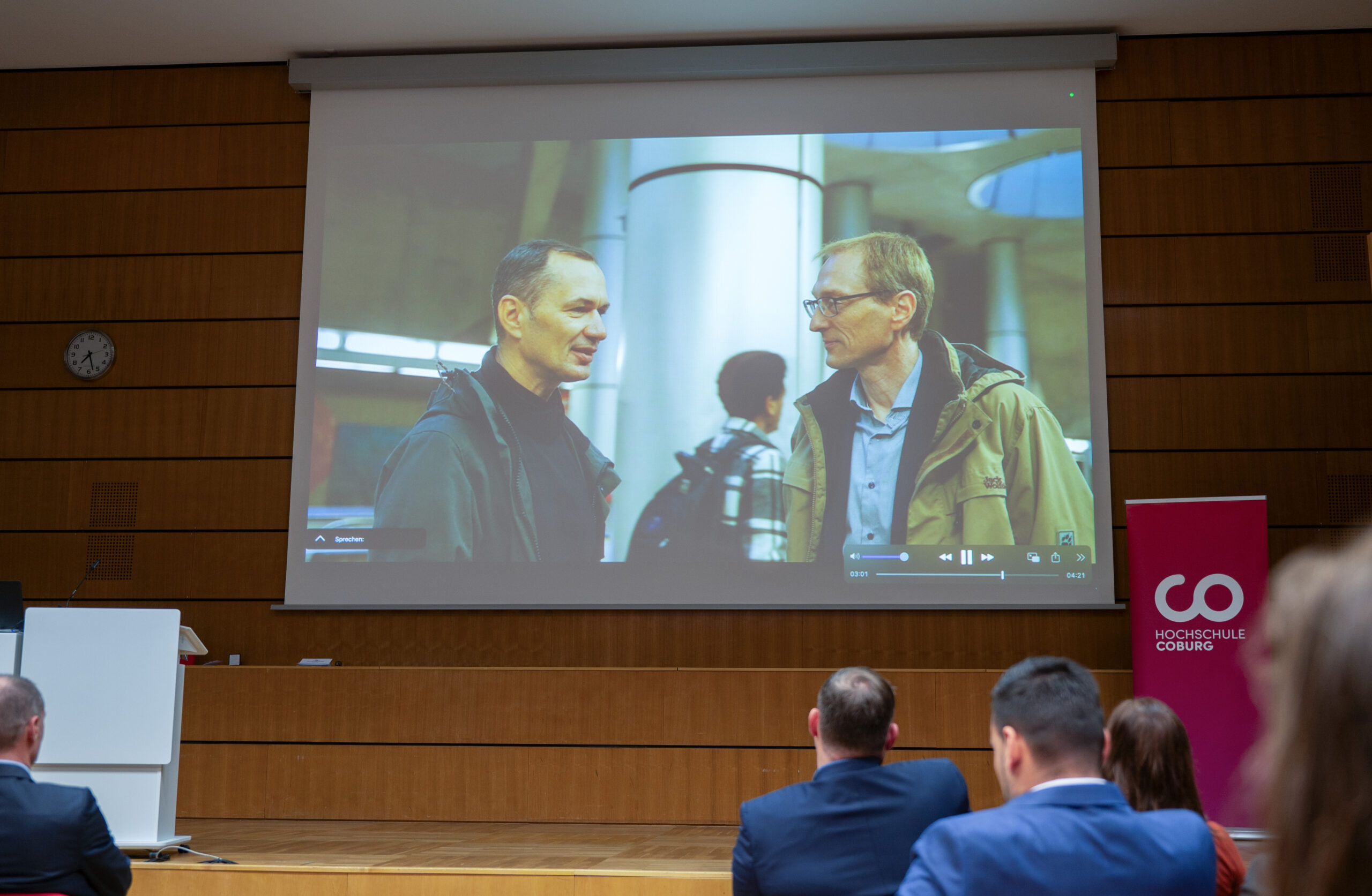 In einem Hörsaal läuft eine Präsentation. Auf einer großen Leinwand sind zwei Männer im Gespräch zu sehen. Die Teilnehmer sitzen und schauen aufmerksam zu. Auf einem roten Banner rechts prangt stolz die Aufschrift „Hochschule Coburg“.