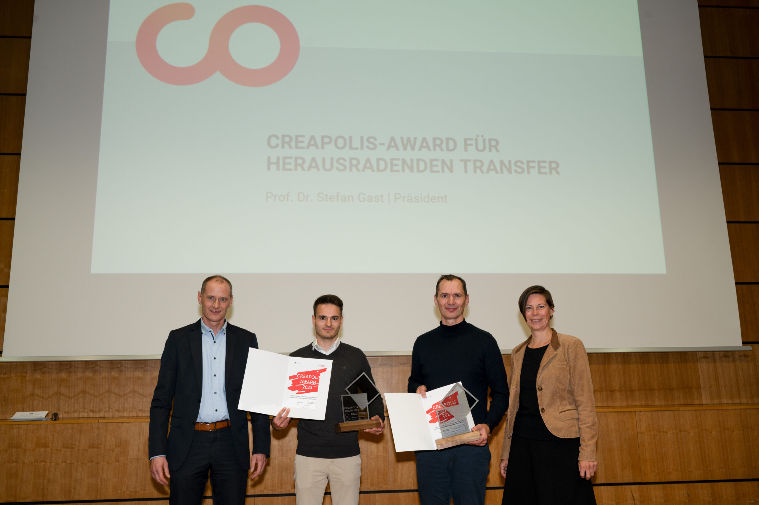 Vier Personen stehen in einer Reihe, zwei halten Urkunden und eine Trophäe in den Händen, vor einer Leinwand mit dem Titel „CREAPOLIS-AWARD FÜR HERAUSRAGENDEN TRANSFER“. Der Rahmen, vermutlich an der Hochschule Coburg, strahlt das Prestige einer Preisverleihung aus.