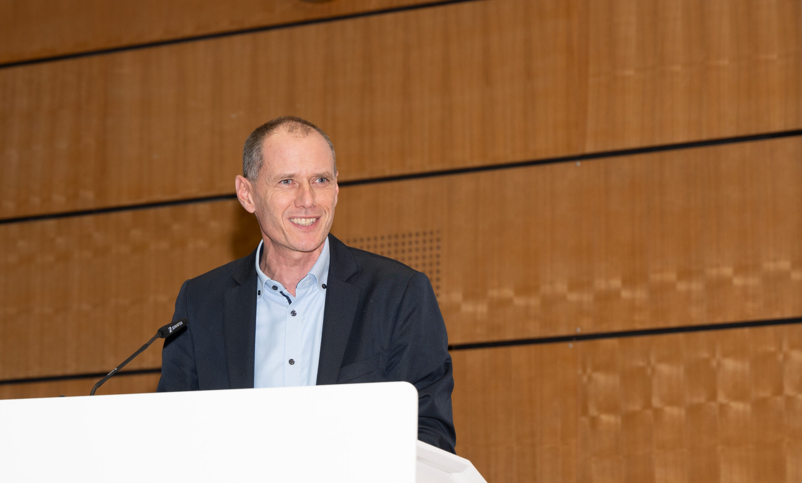 Ein Mann im Anzug spricht an einem Podium in einem großen, holzgetäfelten Raum der Hochschule Coburg. Er lächelt und blickt zur Seite, vor ihm steht ein Mikrofon.