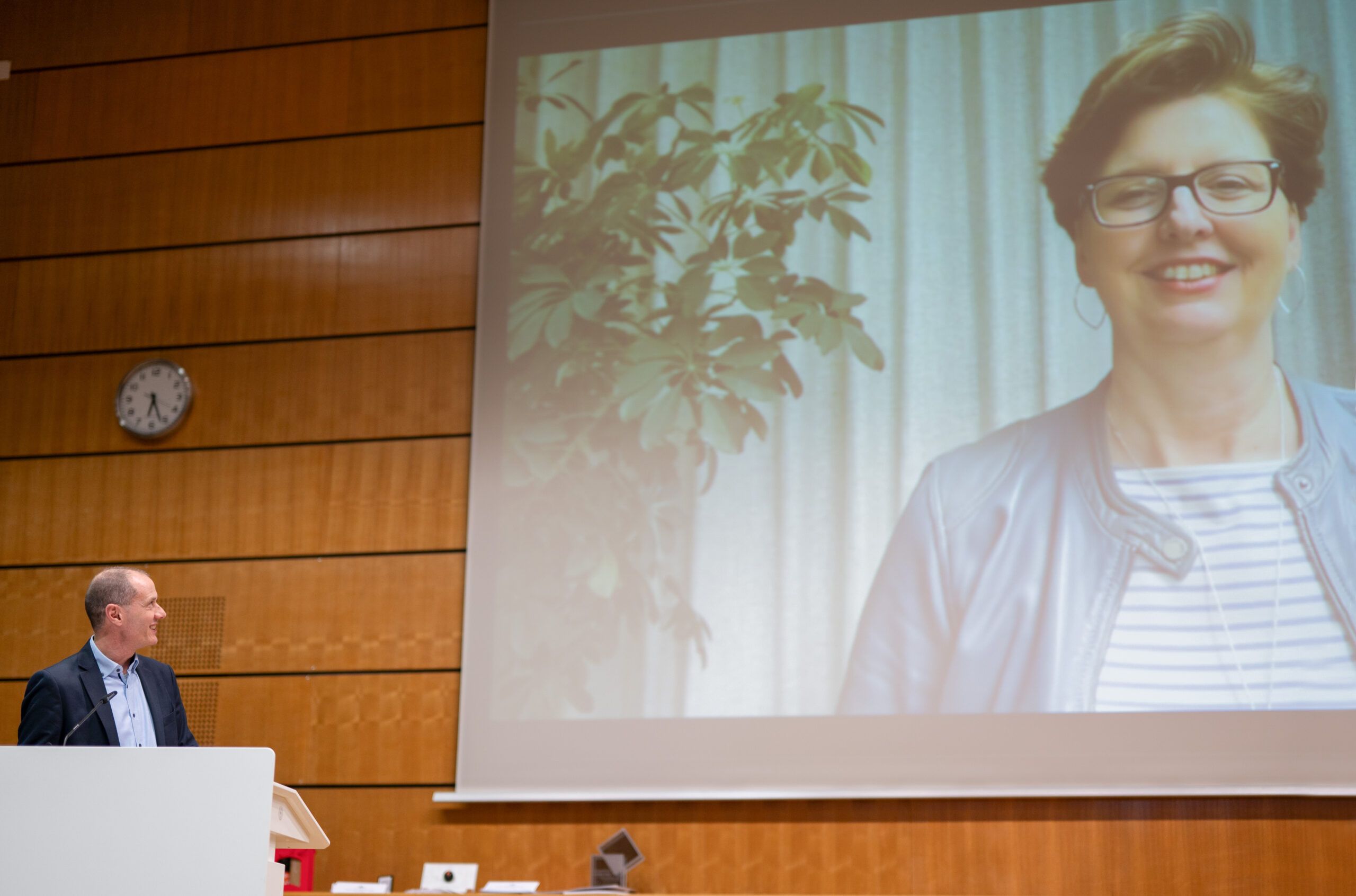 Ein Mann steht an einem Rednerpult in einem Hörsaal der Hochschule Coburg und blickt auf ein großes projiziertes Bild einer lächelnden Frau mit kurzen Haaren und Brille. An der Wand hinter ihm hängt eine Uhr.