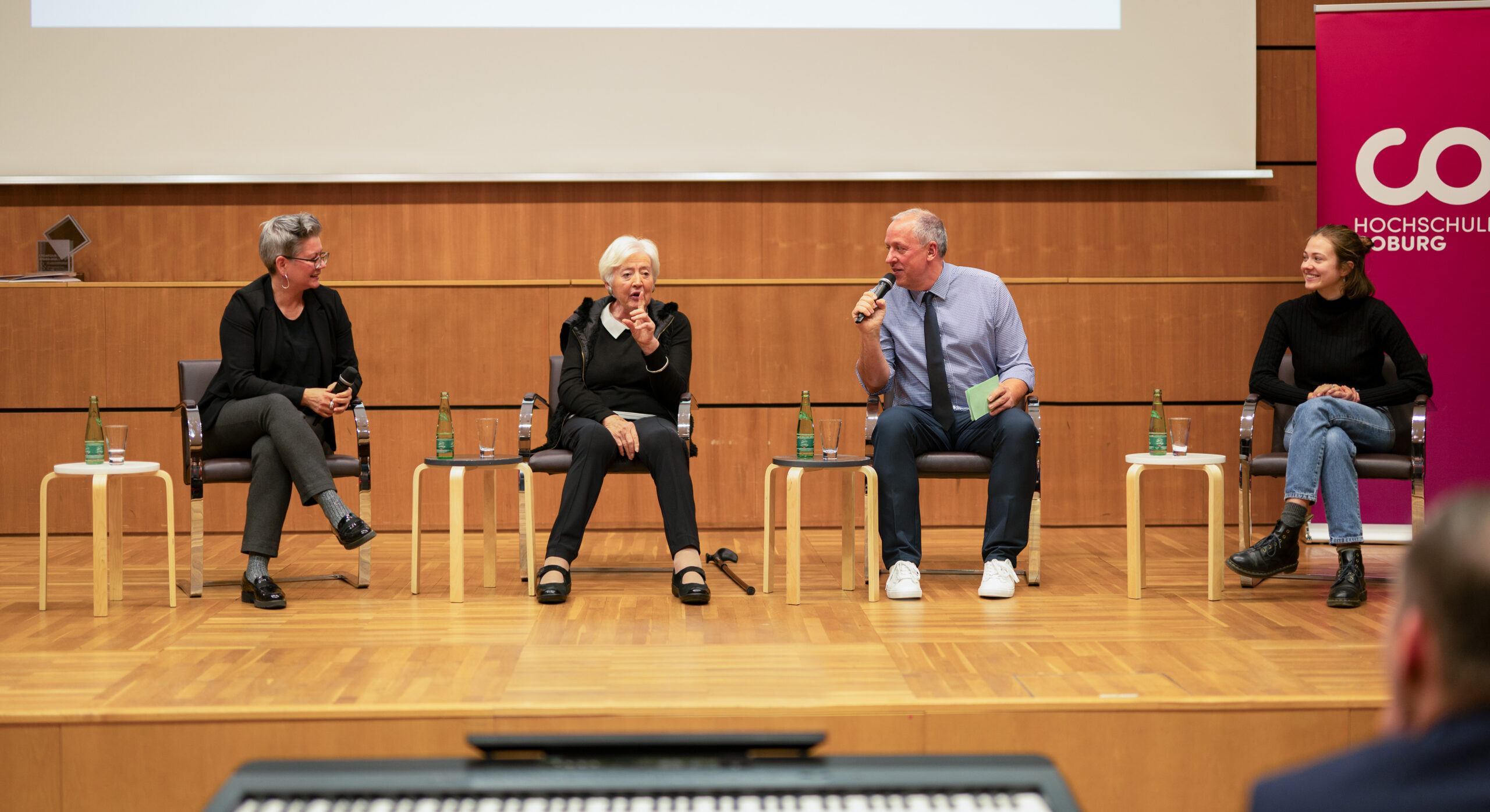 Vier Personen sitzen auf einer Bühne der Hochschule Coburg bei einer Podiumsdiskussion. Sie halten jeweils ein Mikrofon in der Hand und unterhalten sich. Eine hölzerne Kulisse und Tische mit Wasserflaschen bereichern die Szenerie, während ein prominentes Banner der Hochschule Coburg den Hintergrund ziert.