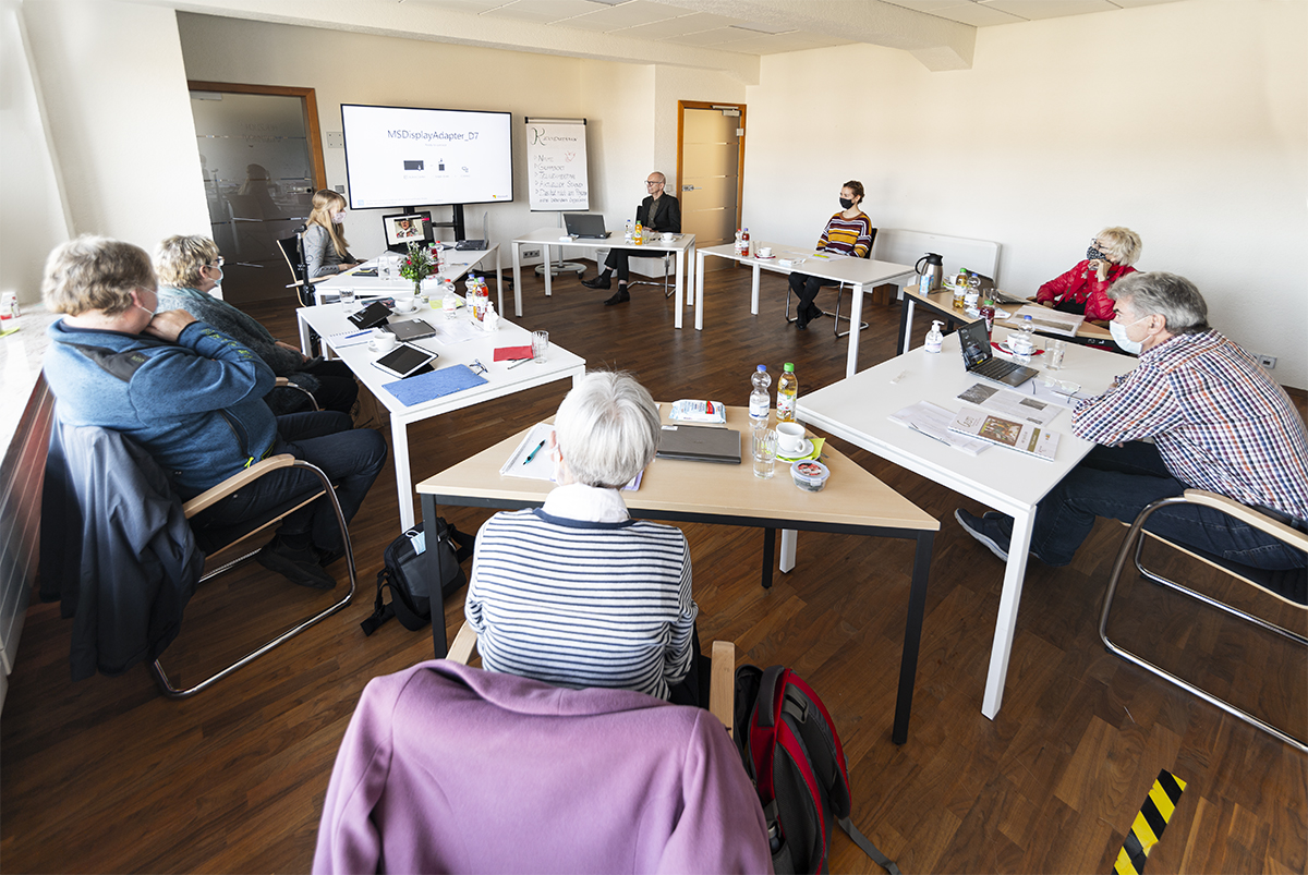 Eine Gruppe von Menschen sitzt um einen U-förmigen Tisch in einem Konferenzraum der Hochschule Coburg und verfolgt aufmerksam eine Präsentation auf dem Bildschirm. Auf den Tischen sind Papiere, Getränke und Laptops verstreut, während ein Vortragender vorne im Raum die Diskussion leitet.