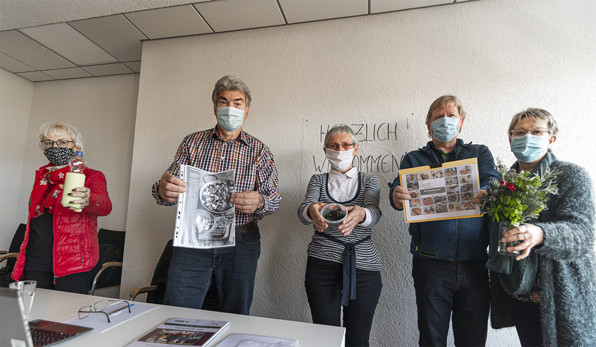 Eine Gruppe von fünf Personen mit Gesichtsmasken steht in einem Gebäude der Hochschule Coburg und hält verschiedene Gegenstände wie Zeitungen, einen Topf und einen Blumenstrauß in den Händen. Sie blicken in die Kamera, an der Wand hinter ihnen hängt ein Schild mit der Aufschrift „Herzlich Willkommen“, das den herzlichen Empfang in der akademischen Umgebung einfängt.