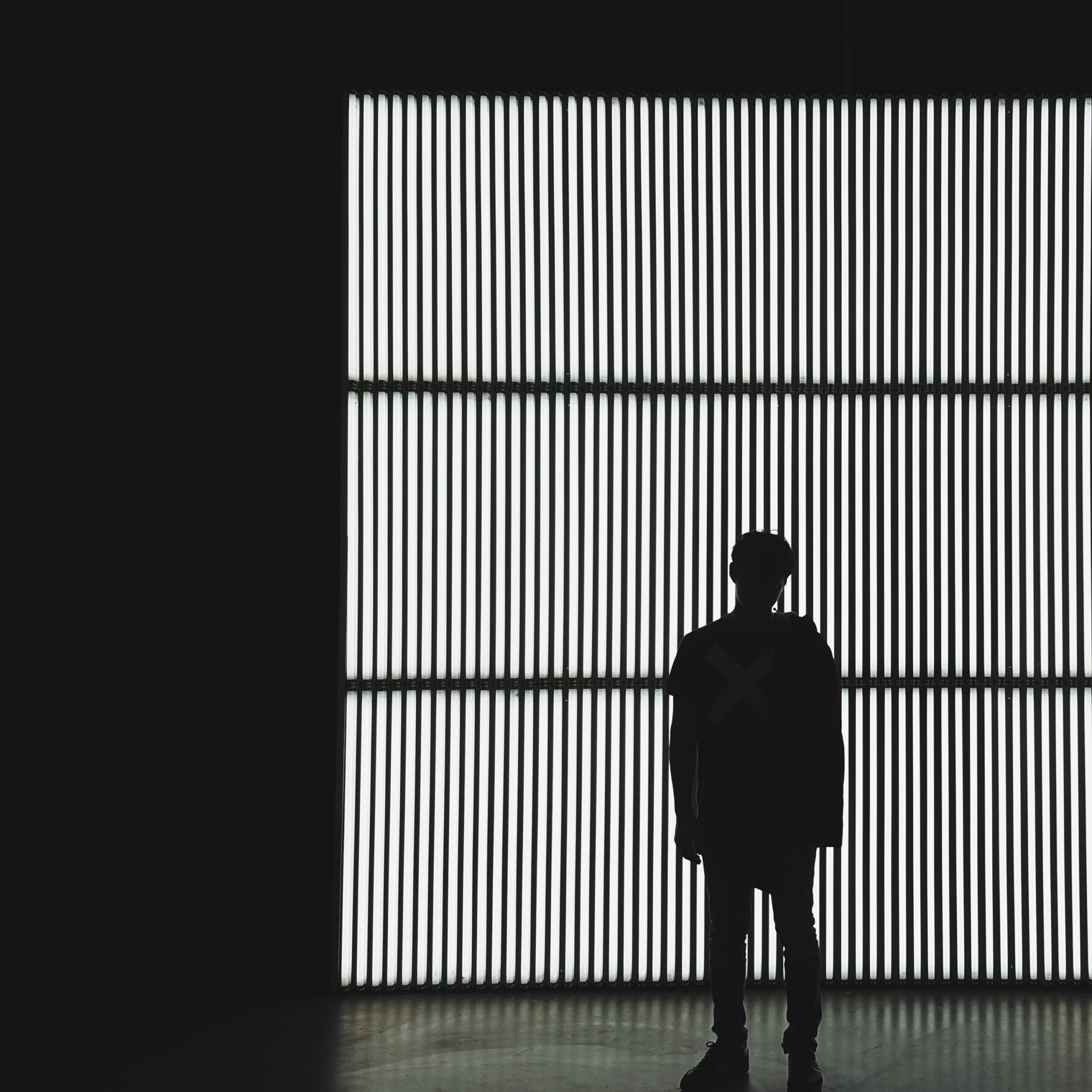 Silhouette einer Person, die vor einem großen, vertikal gestreiften Lichtpaneel in der Hochschule Coburg steht. Im dunklen Raum erzeugt das Licht einen starken Kontrast und wirft Schatten auf die Figur vor dem hellen Hintergrund.