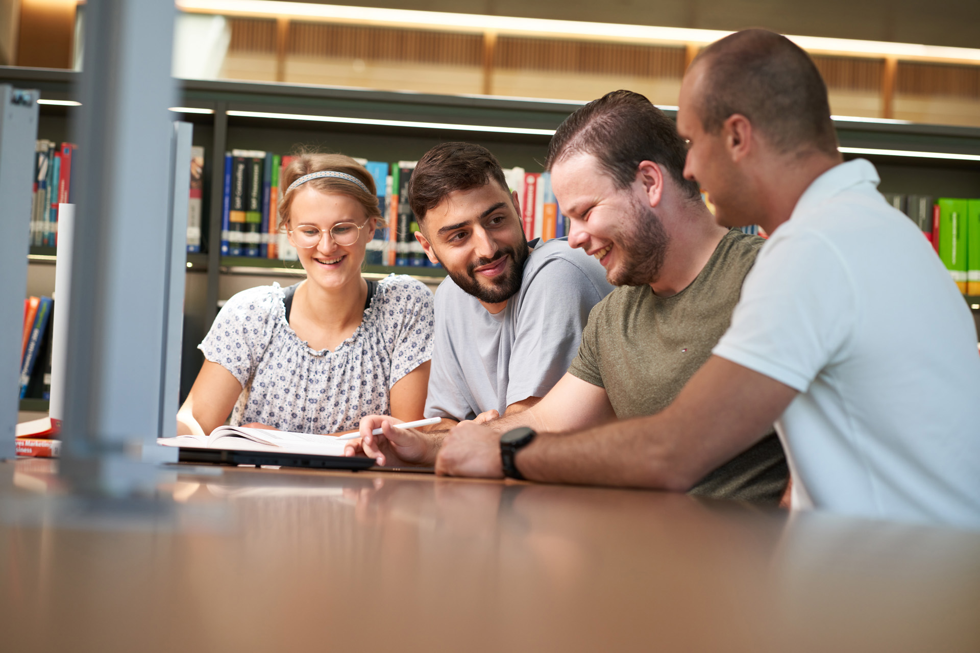 Vier Personen sitzen an einem Tisch in der Bibliothek der Hochschule Coburg, umgeben von Bücherregalen. Sie lächeln und unterhalten sich, vor ihnen liegt ein aufgeklappter Laptop. Die Umgebung wirkt hell und lädt zum Lernen oder Zusammenarbeiten ein.