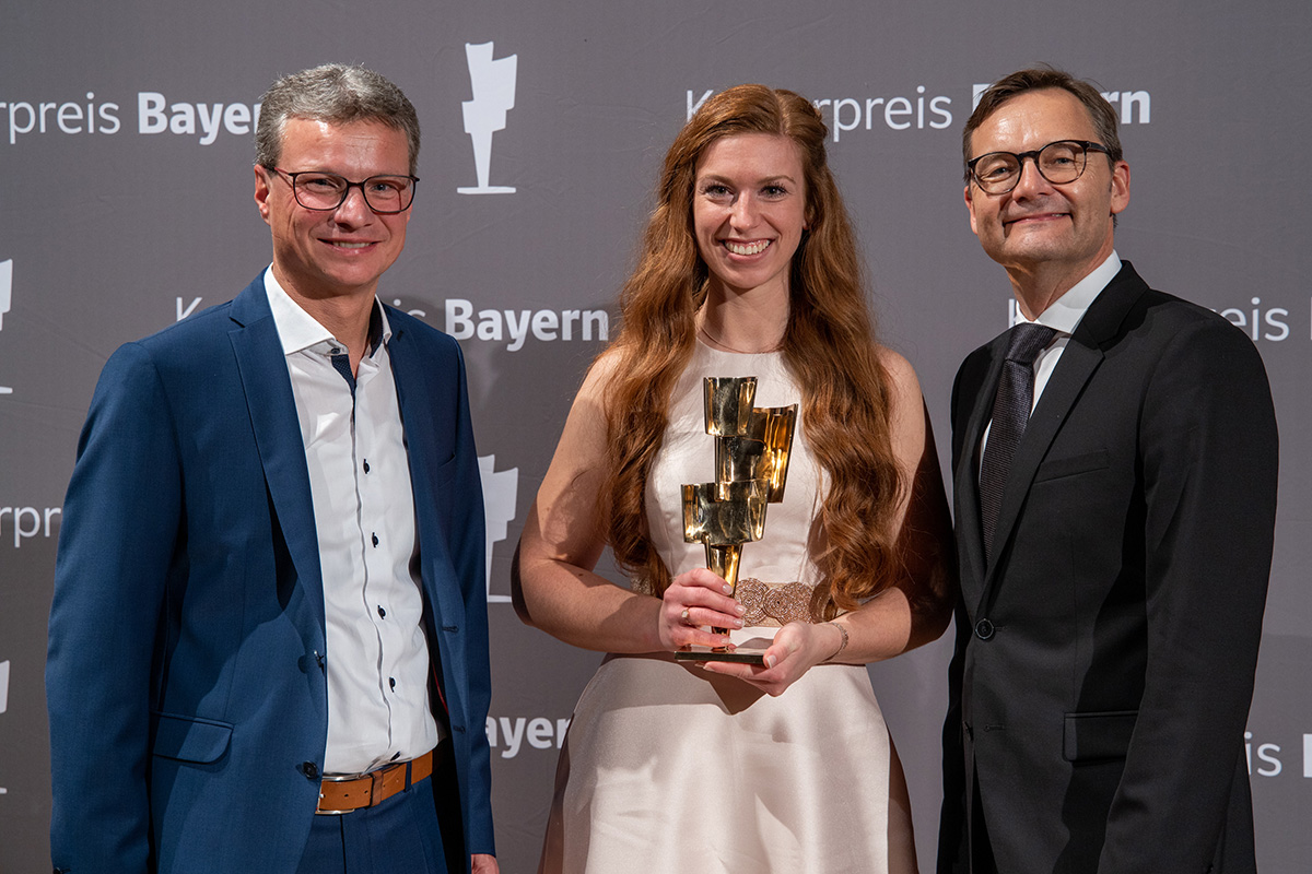 Drei Menschen stehen lächelnd bei einer Preisverleihung zusammen. Die Frau in der Mitte, Vertreterin der Hochschule Coburg, hält eine Trophäe in der Hand. Sie sind in formelle Kleidung gekleidet und stehen vor einem Hintergrund mit der Aufschrift „Kulturpreis Bayern“.