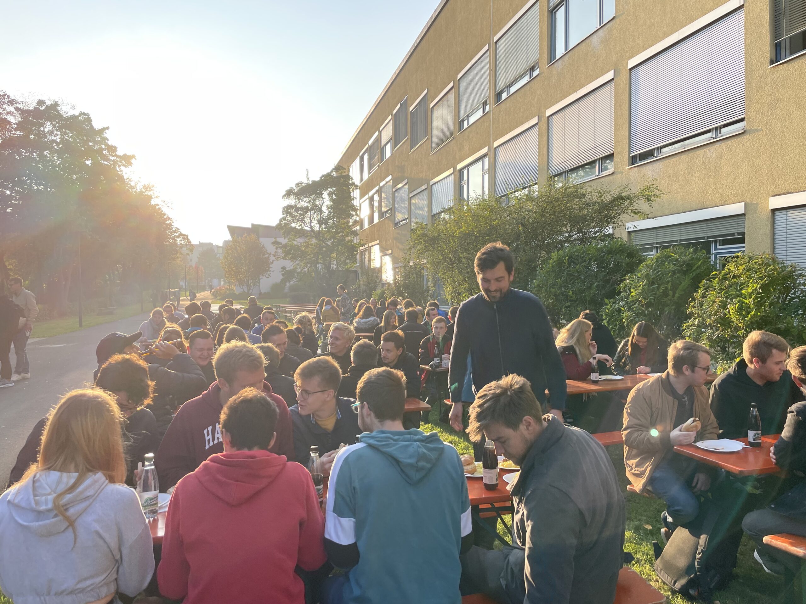 Eine große Gruppe von Menschen sitzt an langen orangefarbenen Tischen vor einem Gebäude der Hochschule Coburg und genießt einen sonnigen Tag. Sie scheinen sich zu unterhalten und zu essen, mit Bäumen und Sonnenlicht im Hintergrund.