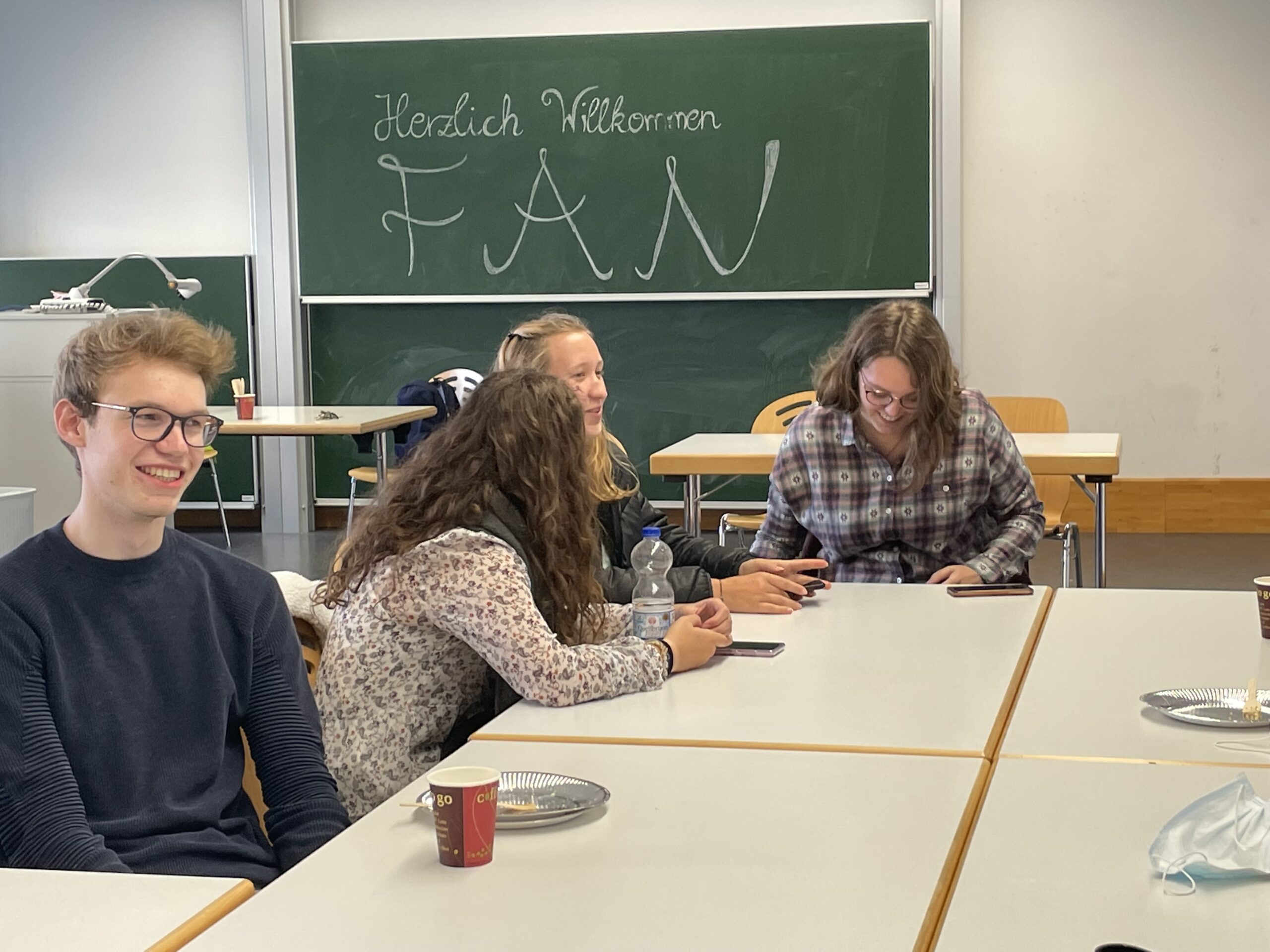 Eine Gruppe von vier Personen sitzt an Tischen in einem Hörsaal, vor ihnen stehen Tassen und Teller. Im Hintergrund steht auf einer Tafel „Herzlich Willkommen FAN, Hochschule Coburg“. Alle scheinen in ein Gespräch vertieft zu sein.