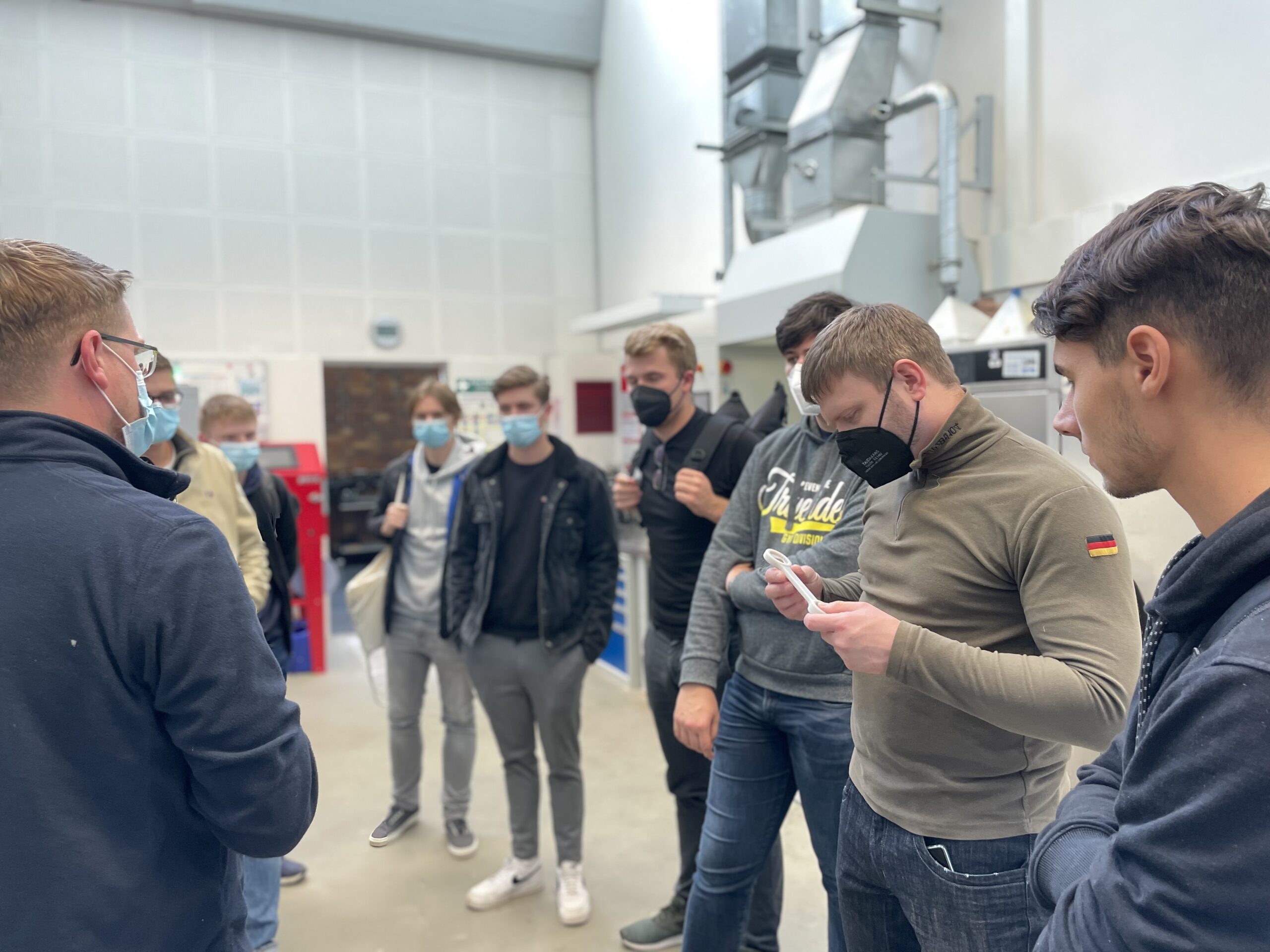 Eine Gruppe von Menschen mit Mund-Nasen-Schutz steht in einer Werkstatt der Hochschule Coburg. Sie unterhalten sich, eine Person hält einen Gegenstand in der Hand, die anderen schauen aufmerksam zu. Im Hintergrund sind verschiedene Werkstattgeräte und Maschinen zu sehen.