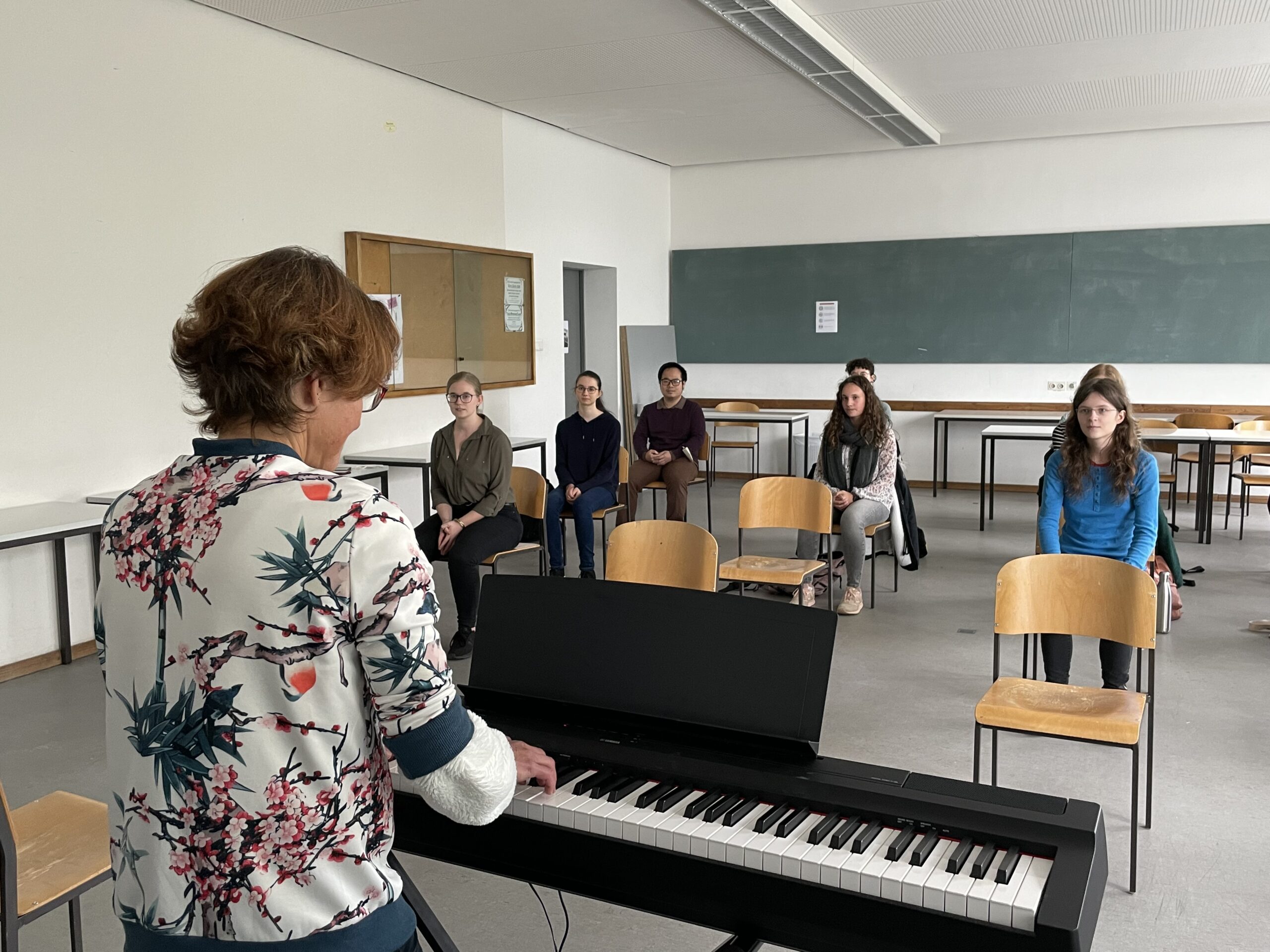 In einem ungezwungenen Unterrichtsraum an der Hochschule Coburg spielt eine Person Keyboard, während fünf andere aufmerksam dasitzen, umgeben von leeren Stühlen, einer großen Tafel und einem Whiteboard. Die Szene lässt auf eine vertrauliche Gruppenzusammenkunft oder Unterrichtsstunde schließen.