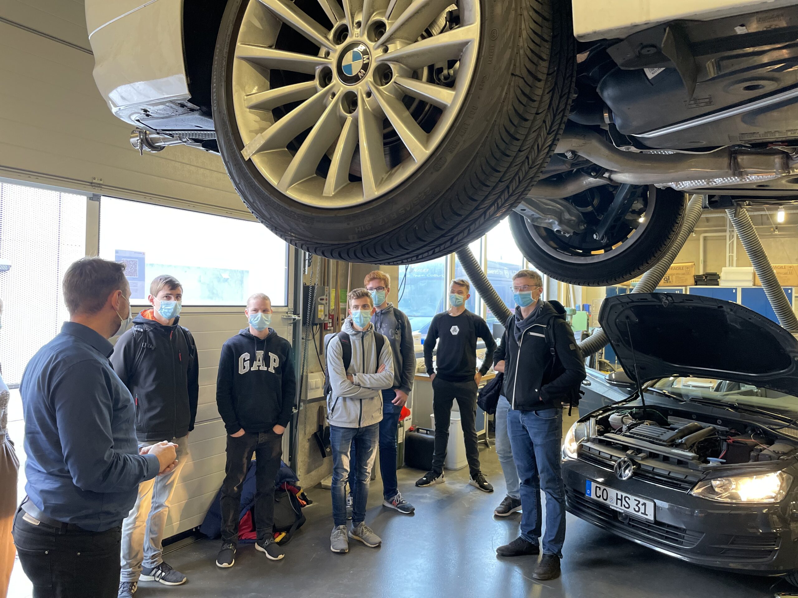 Eine Gruppe maskierter Menschen steht in einer Autowerkstatt der Hochschule Coburg. Ein Auto ist auf einer Hebebühne hochgehoben, so dass sein Unterboden sichtbar ist, während bei einem anderen Auto die Motorhaube geöffnet ist. Eine Person scheint der Gruppe etwas zu erklären oder Anweisungen zu geben.