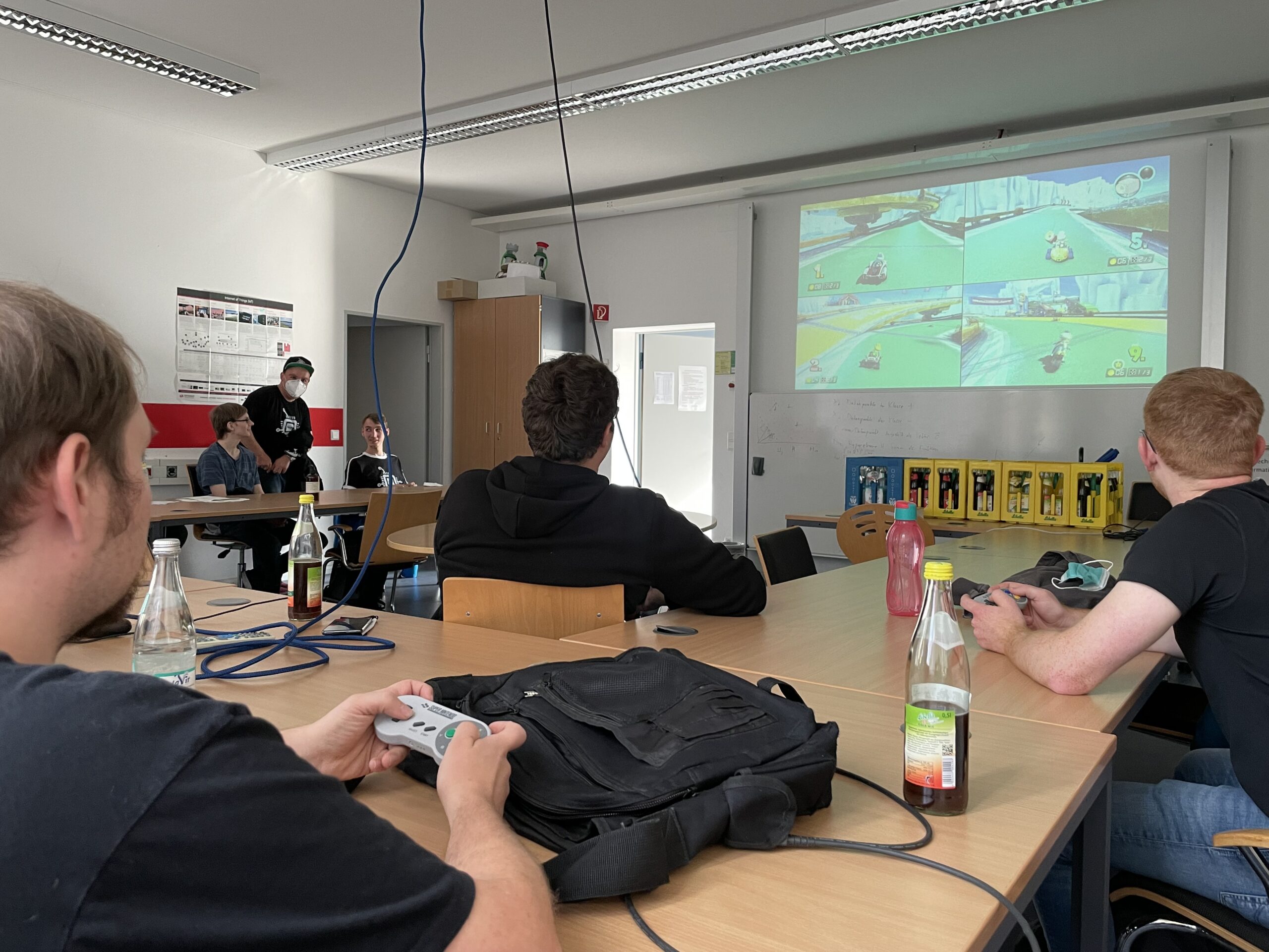 Eine Gruppe von Menschen sitzt an einem Tisch in der Hochschule Coburg und spielt ein Multiplayer-Videospiel, das an die Wand projiziert wird. In diesem gemütlich beleuchteten Raum sind Gamecontroller im Einsatz und verschiedene Getränke auf den Büromöbeln verteilt.