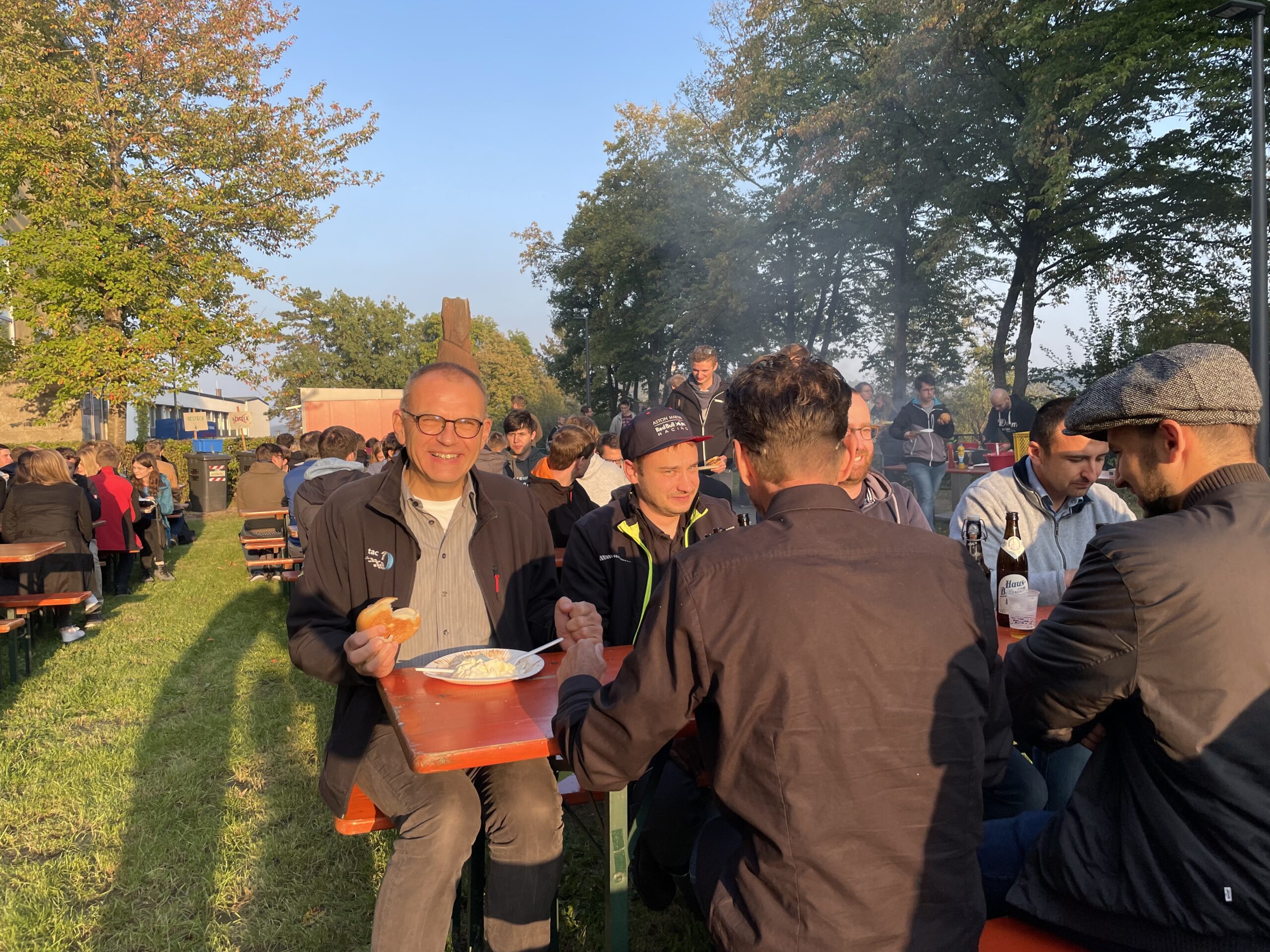 Eine Gruppe von Menschen der Hochschule Coburg sitzt an einem sonnigen Tag an Picknicktischen in einem Park. Eine Person im Vordergrund lächelt, während sie aus einer Schüssel isst, während andere Essen und Getränke genießen. Im Hintergrund sind Bäume und weitere Menschen zu sehen, die zur lebhaften Atmosphäre beitragen.