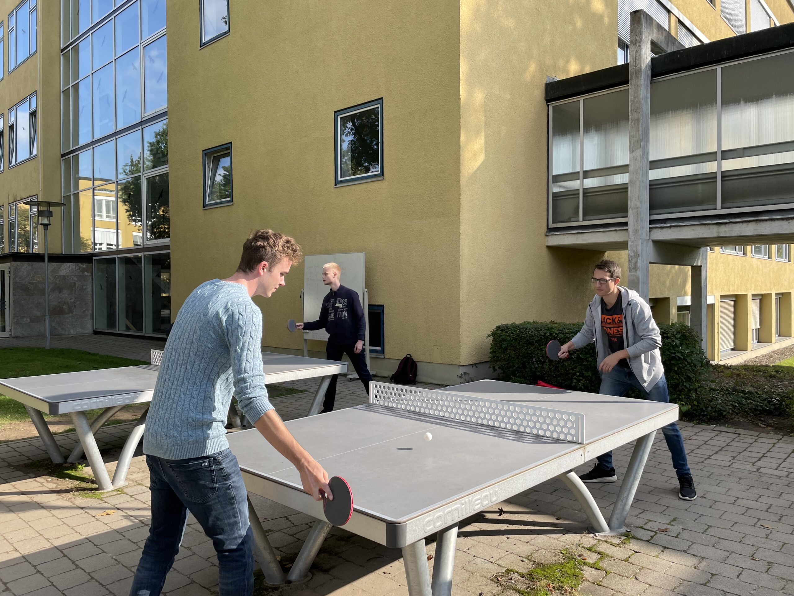 Drei Personen spielen an einem sonnigen Tag im Freien in der Nähe der Hochschule Coburg eine Partie Tischtennis. Zwei Spieler schwingen hochkonzentriert ihre Schläger, während der Ball zwischen ihnen hin und her saust, während der dritte von neben einem gelben Gebäude mit großen Fenstern zusieht.