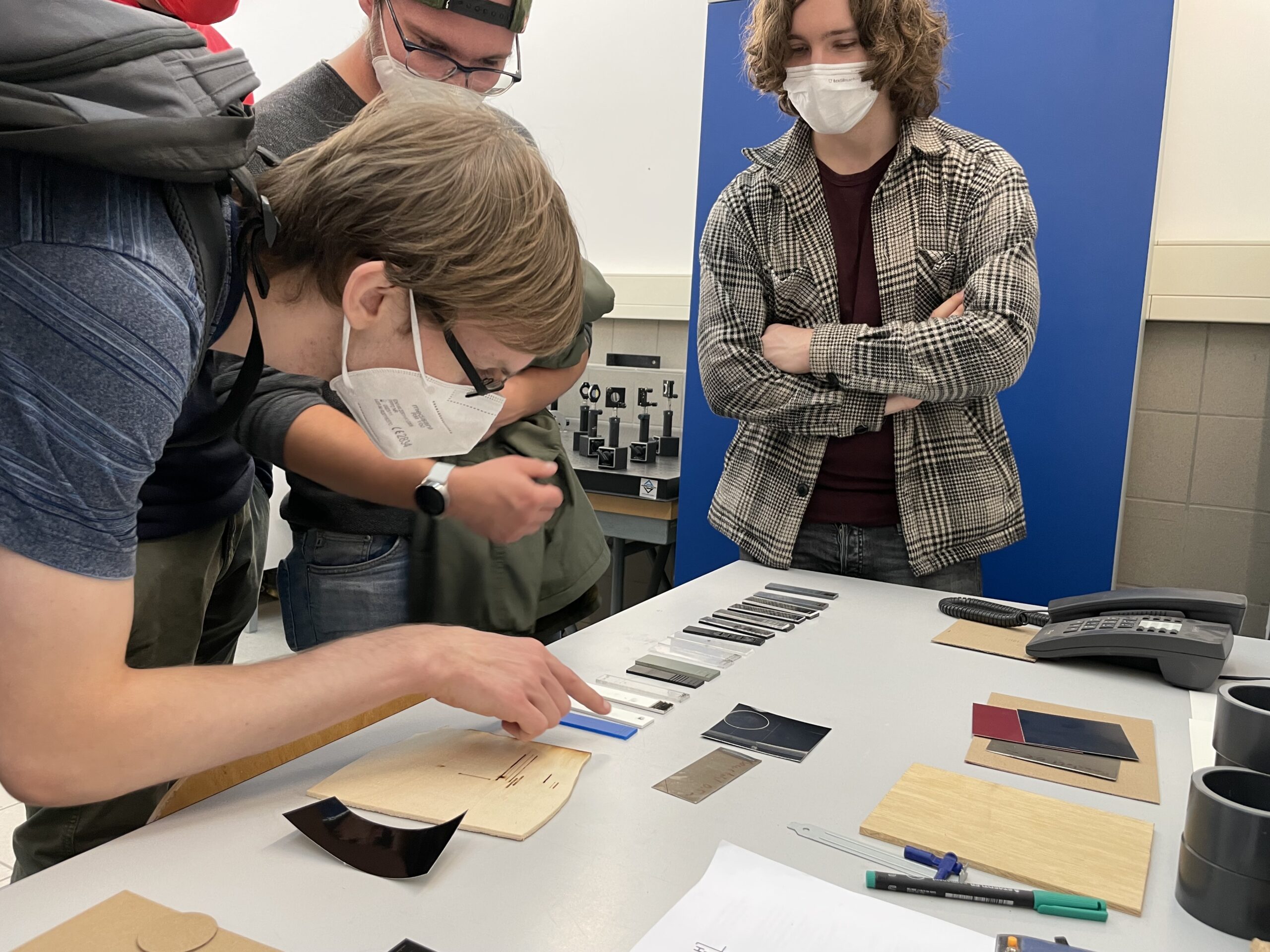 Vier Personen befinden sich in einem Raum der Hochschule Coburg und untersuchen Materialien auf einem Tisch, darunter verschiedene Proben wie Fliesen und Platten. Zwei Personen tragen Masken, während bei der kreativen Erkundung auch ein Telefon und einige Dokumente zu sehen sind.