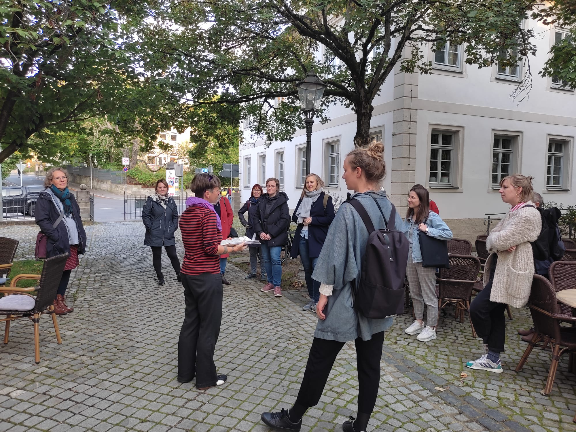 Eine Gruppe von Menschen steht draußen in einem gepflasterten Innenhof, umgeben von Bäumen und Gebäuden der Hochschule Coburg. Eine Person spricht zu der Gruppe, während andere aufmerksam zuhören. Einige halten Taschen und tragen Jacken. In der Nähe sind Stühle und ein Tisch zu sehen.