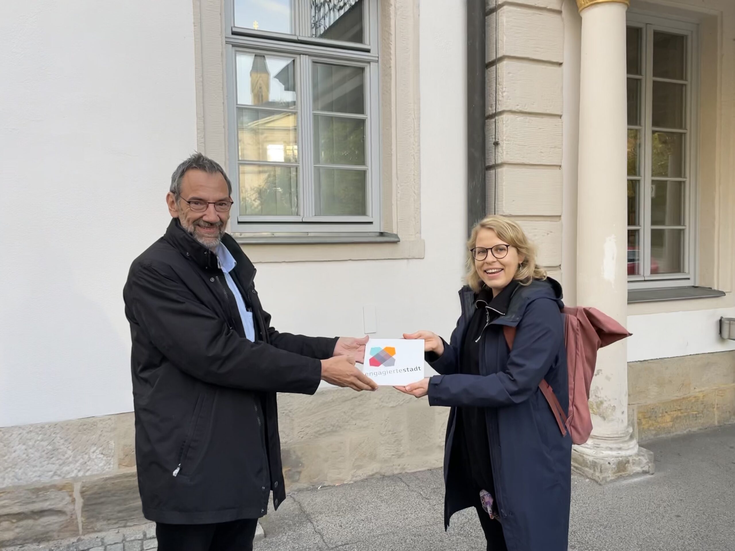 Zwei Menschen lächeln und halten gemeinsam eine weiße Auszeichnungsplakette vor einem Gebäude mit Säulen und Bogenfenstern der Hochschule Coburg. Die Person links trägt einen schwarzen Mantel, die Person rechts trägt eine Brille, eine dunkle Jacke und einen roten Rucksack.