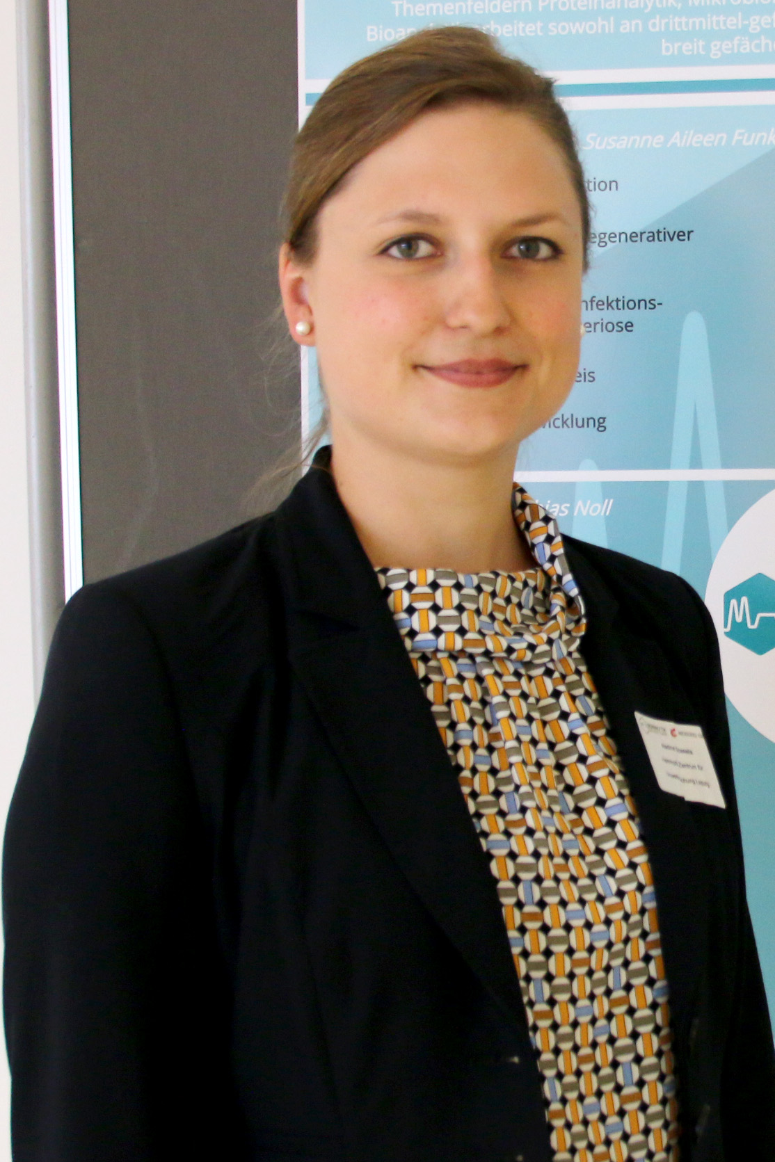 Eine Person mit hellbraunem Haar, einem schwarzen Blazer und einer gemusterten Bluse steht selbstbewusst vor einer Tafel mit Text an der Hochschule Coburg.