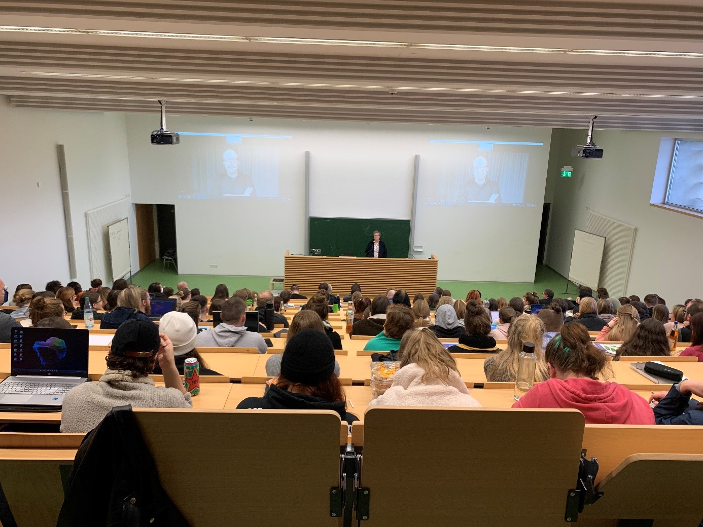 In einem überfüllten Hörsaal der Hochschule Coburg herrscht reges Treiben. Studenten sitzen vor einem Dozenten. Der Dozent steht an einem Rednerpult vor einer großen Tafel. Auf zwei Bildschirmen läuft eine Präsentation. Der Raum ist hell erleuchtet, die Holztische stehen in Reihen, die Wände sind weiß.