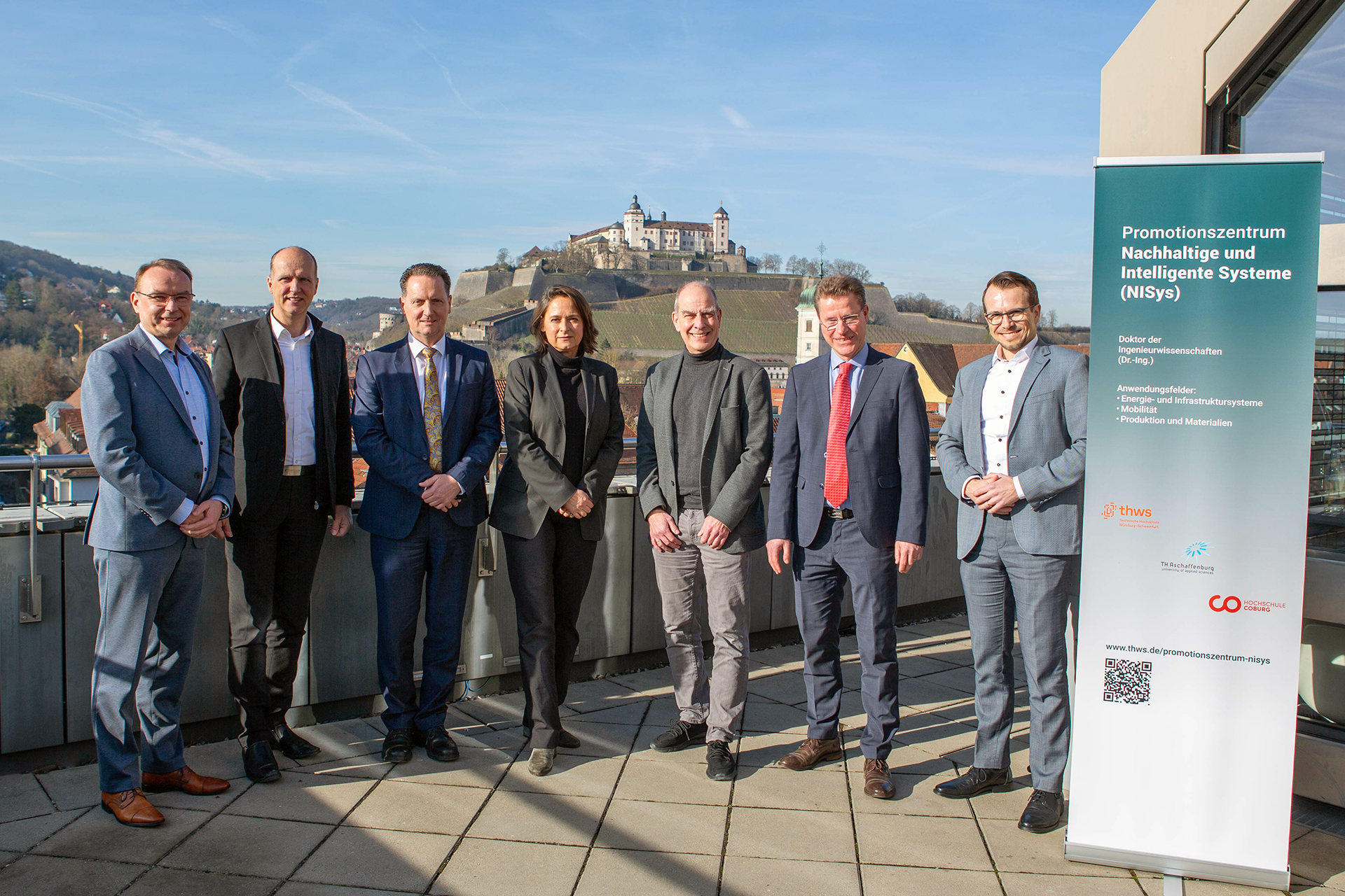 A group of people in formal attire stand on a terrace with a scenic view of a castle on a hill. They pose next to a promotional banner for the Hochschule Coburg's "Promotionszentrum Nachhaltige und Intelligente Systeme (NISys).