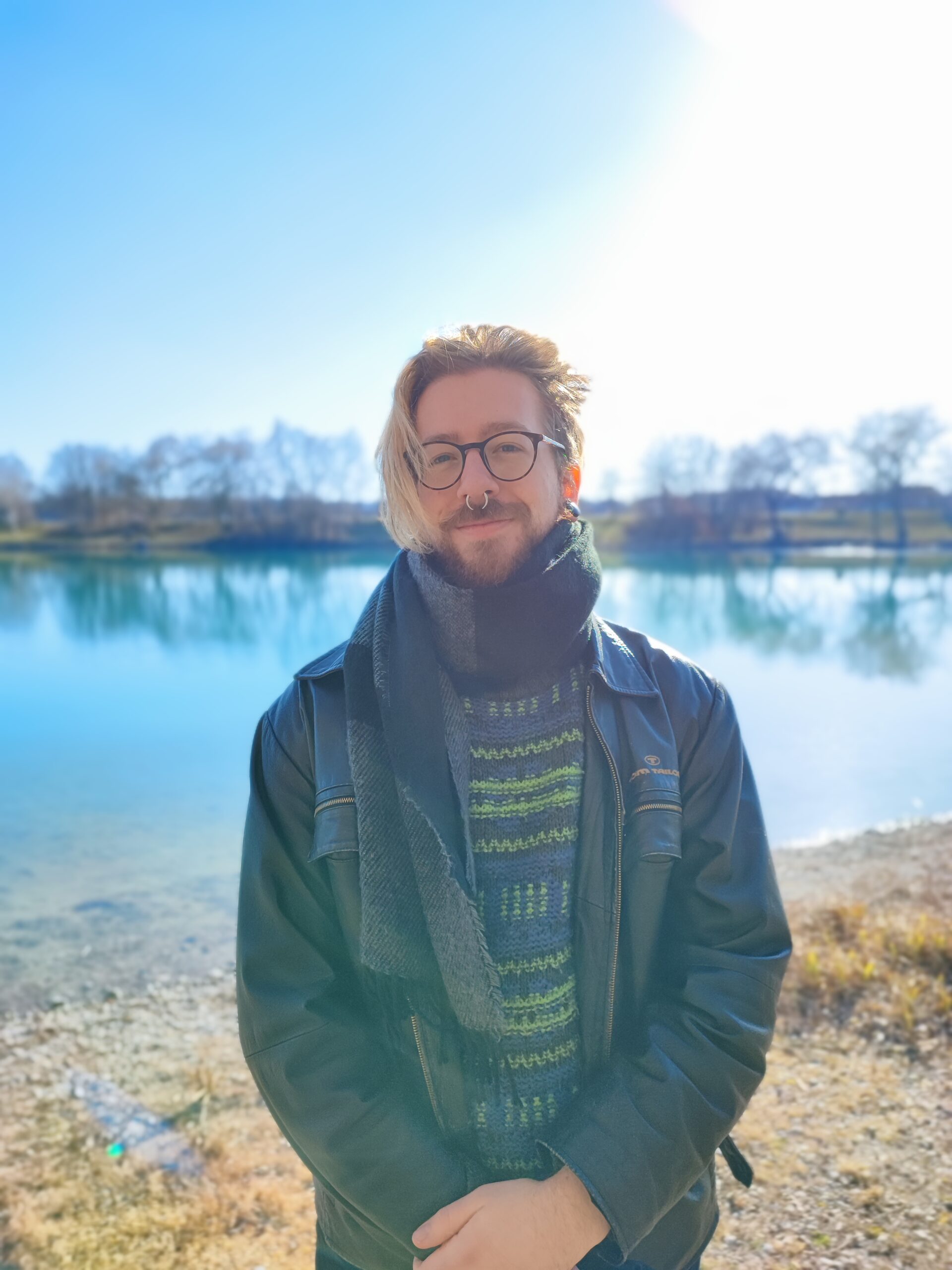 Eine Person mit Brille, Nasenring und Bart steht lächelnd am See in der Nähe der Hochschule Coburg. Sie trägt eine schwarze Lederjacke, einen grün gemusterten Pullover und einen dunklen Schal. Im Hintergrund sind ruhiges Wasser und Bäume unter einem strahlend blauen Himmel zu sehen.