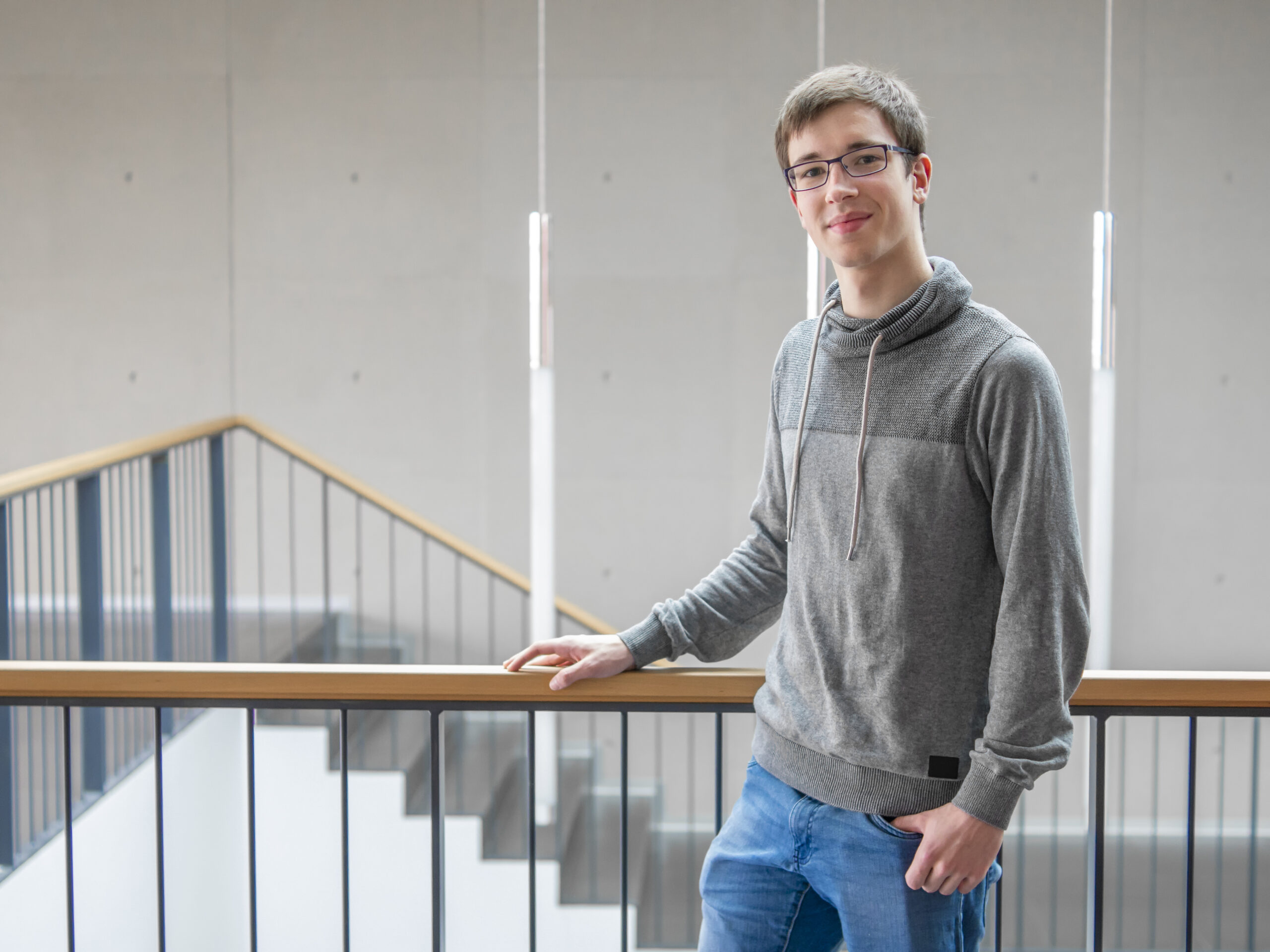 Eine Person mit Brille und grauem Pullover lehnt an einem Geländer in einem modernen Gebäude der Hochschule Coburg, im Hintergrund windet sich eine Treppe elegant.