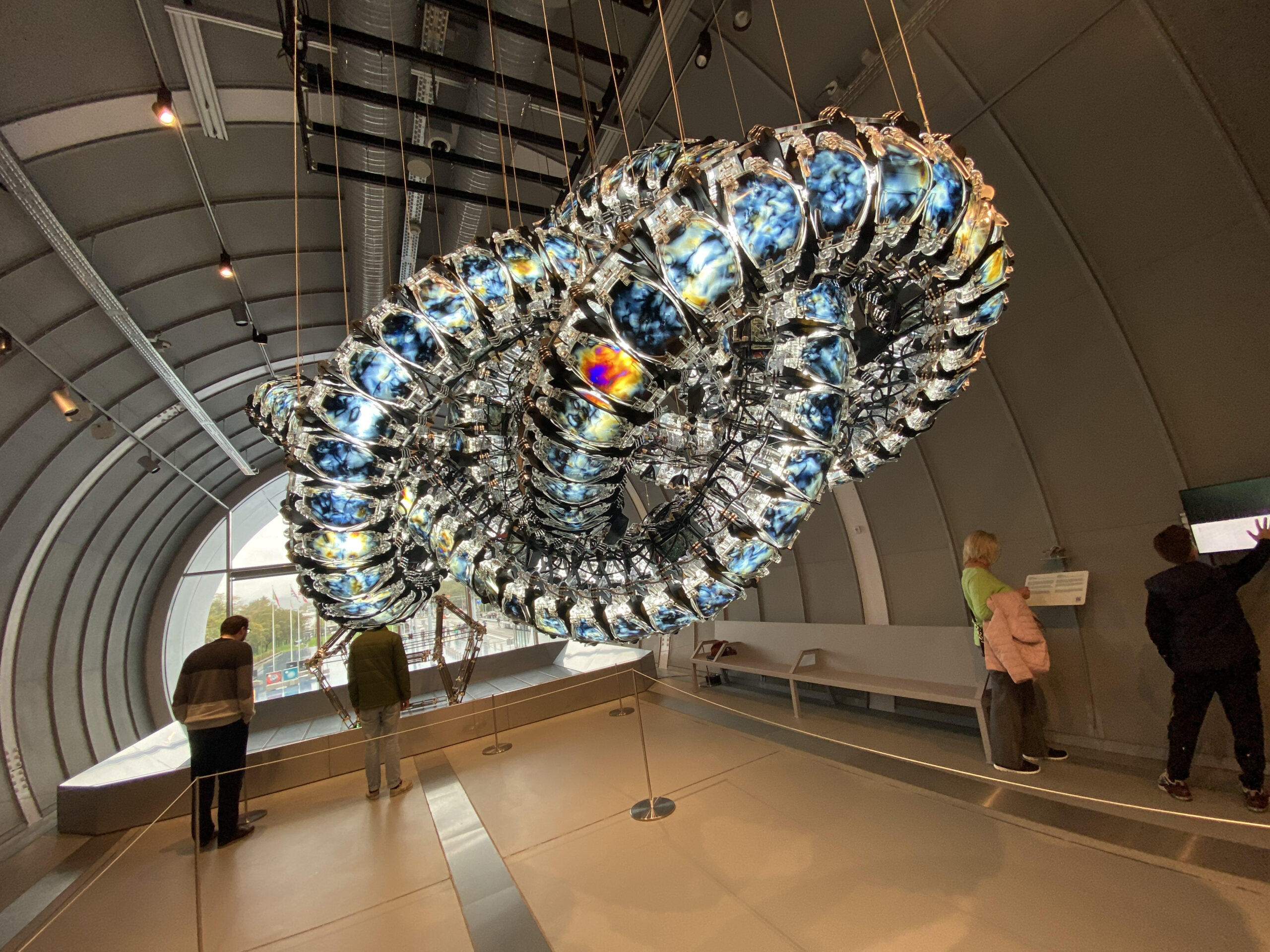 In einer modernen, grauen Galerie der Hochschule Coburg hängt eine große, abstrakte Skulptur von der Decke. Das Kunstwerk besteht aus reflektierenden, metallischen Segmenten mit eingebetteten bunten Bildern. Besucher stehen herum und betrachten die Installation.