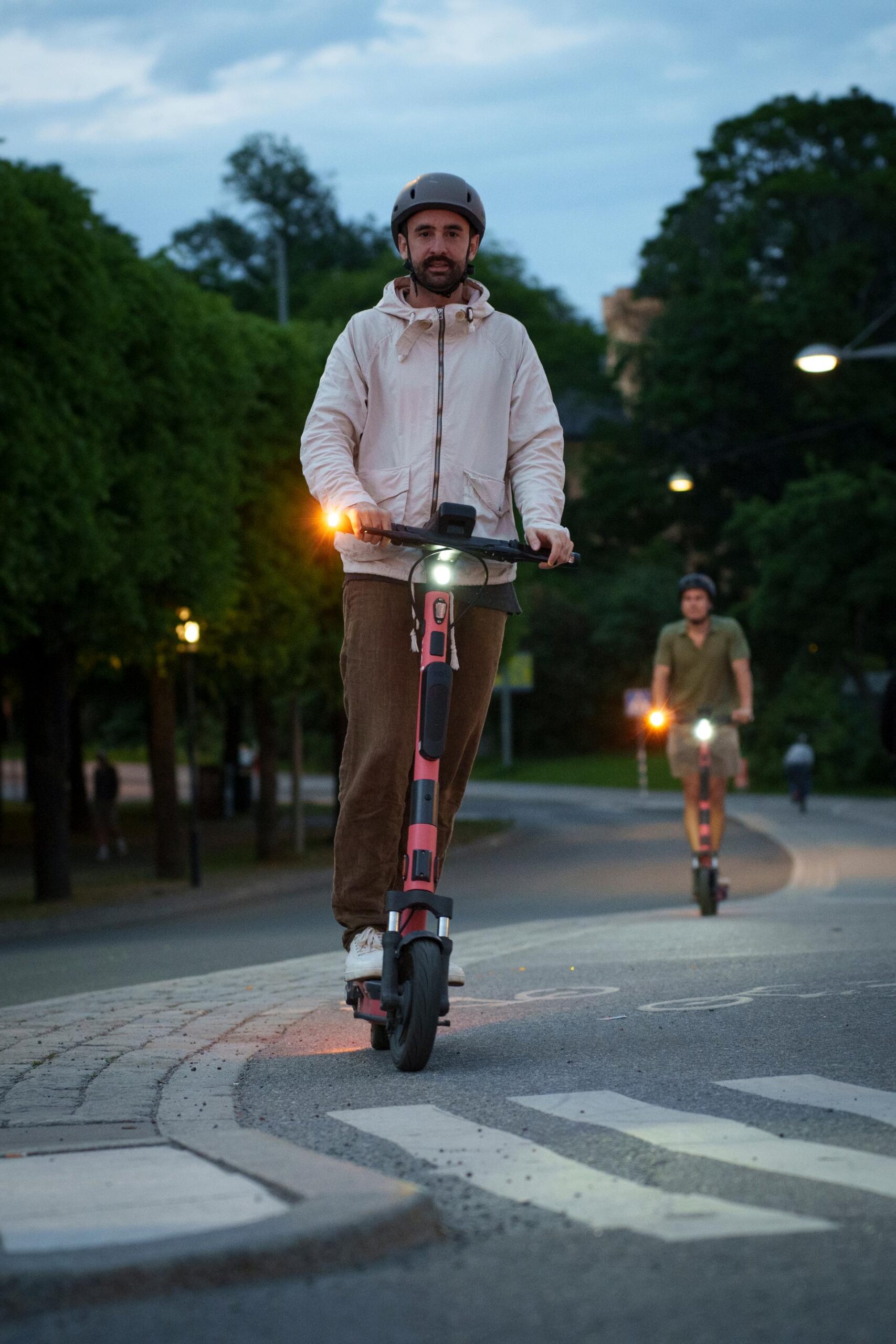 Eine Person mit Helm fährt mit einem Elektroroller auf einer kurvigen, von Bäumen gesäumten Straße in der Nähe der Hochschule Coburg. Im Hintergrund folgt ein weiterer Rollerfahrer. Es ist ein bewölkter Tag und die Roller haben Lichter an.