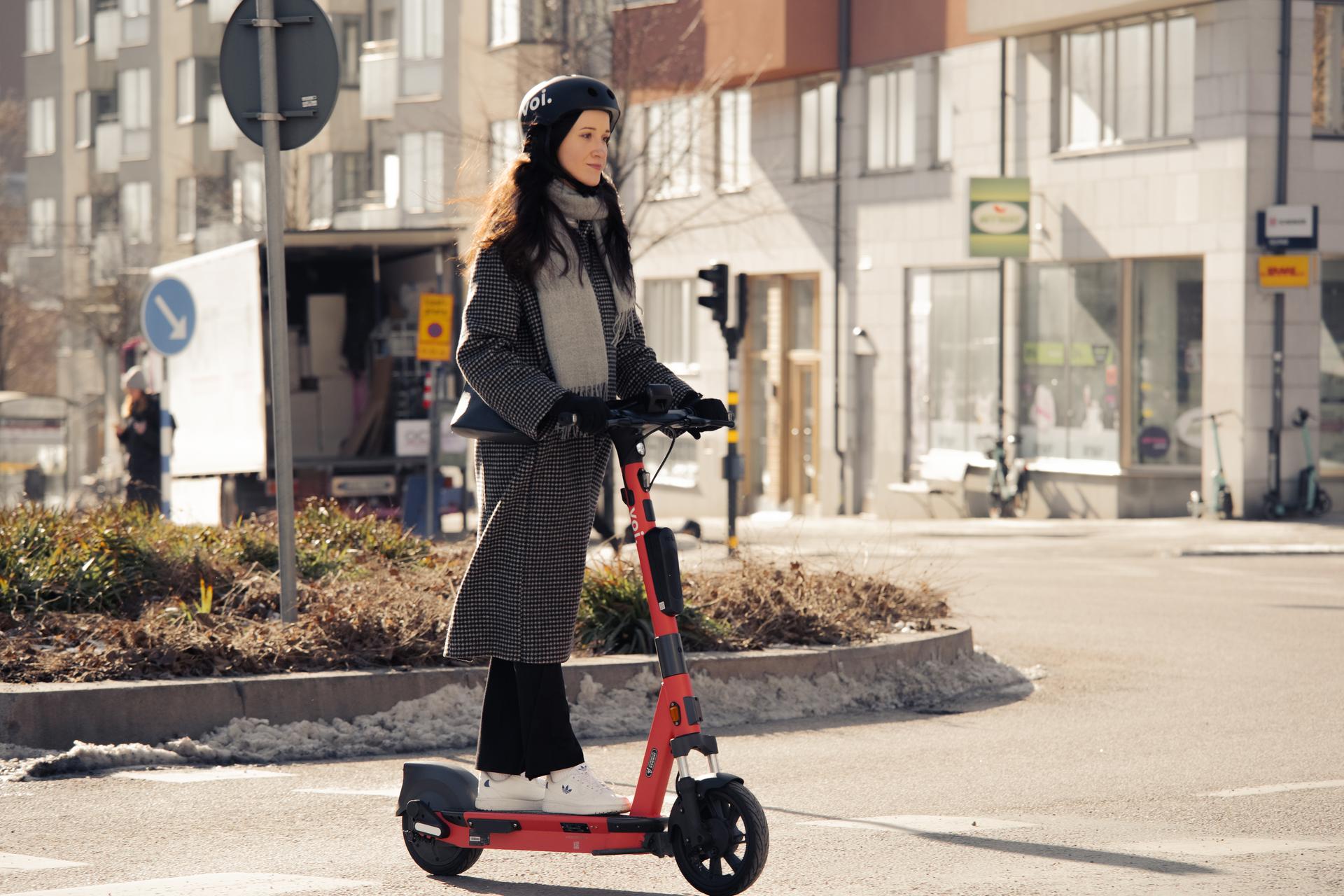 Eine Person mit Helm und Mantel fährt mit einem roten Elektroroller an der Hochschule Coburg auf einer Stadtstraße vorbei. Im Hintergrund sind Gebäude und ein Lieferwagen zu sehen, die Sonne wirft Schatten auf die Straße.