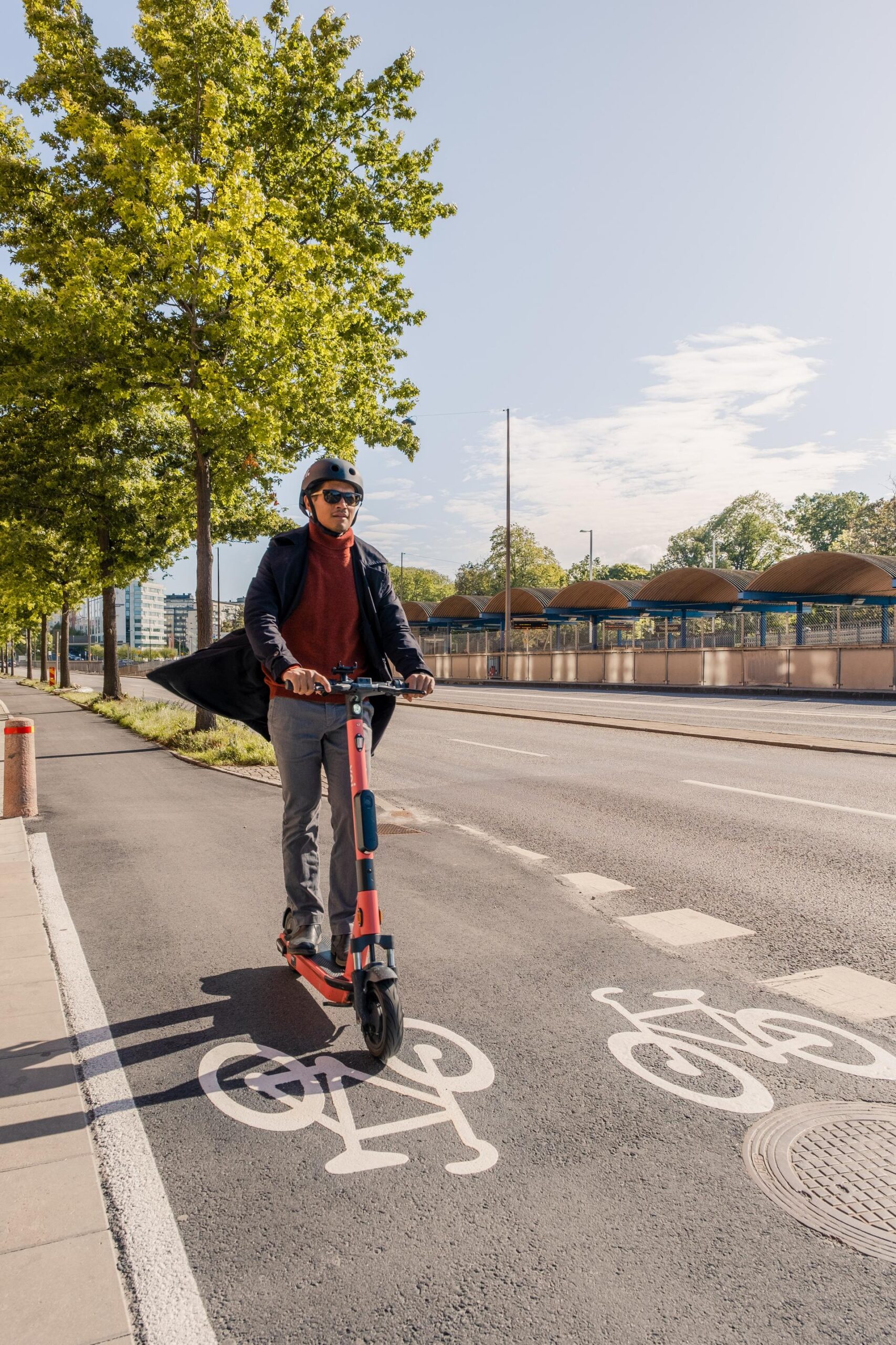 Eine Person fährt an einem sonnigen Tag mit einem Elektroroller auf einem ausgewiesenen Radweg in der Nähe der Hochschule Coburg. Sie trägt einen Helm und eine Sonnenbrille. Eine Reihe von Bäumen säumt die Straße. Im Hintergrund sind ein großes Gebäude und mehrere Bushaltestellen zu sehen.