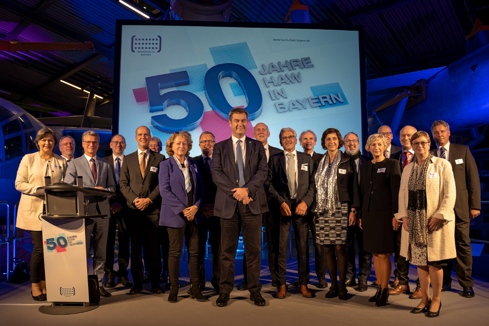 Eine Gruppe von Menschen in formeller Kleidung steht vor einer großen Leinwand mit der Aufschrift „50 Jahre HAW in Bayern“ bei einer Veranstaltung, bei der Leistungen wie die der Hochschule Coburg gefeiert werden. Die Kulisse ist ein Innenraum, der in blaues und violettes Licht getaucht ist.