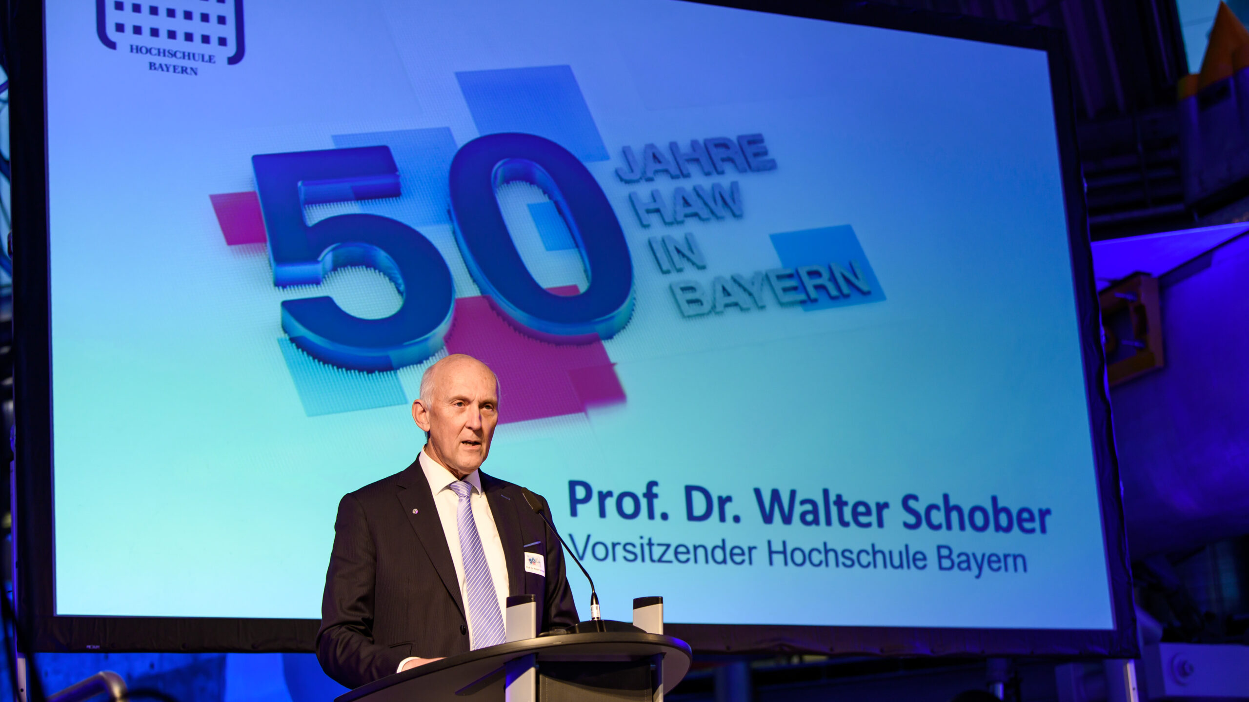 Ein Mann steht auf einem Podium und spricht bei einer Veranstaltung. Hinter ihm zeigt eine große Leinwand „50 Jahre HAW in Bayern“ und den Namen „Prof. Dr. Walter Schober, Vorsitzender Hochschule Bayern“ und würdigt akademische Exzellenz neben Institutionen wie der Hochschule Coburg.