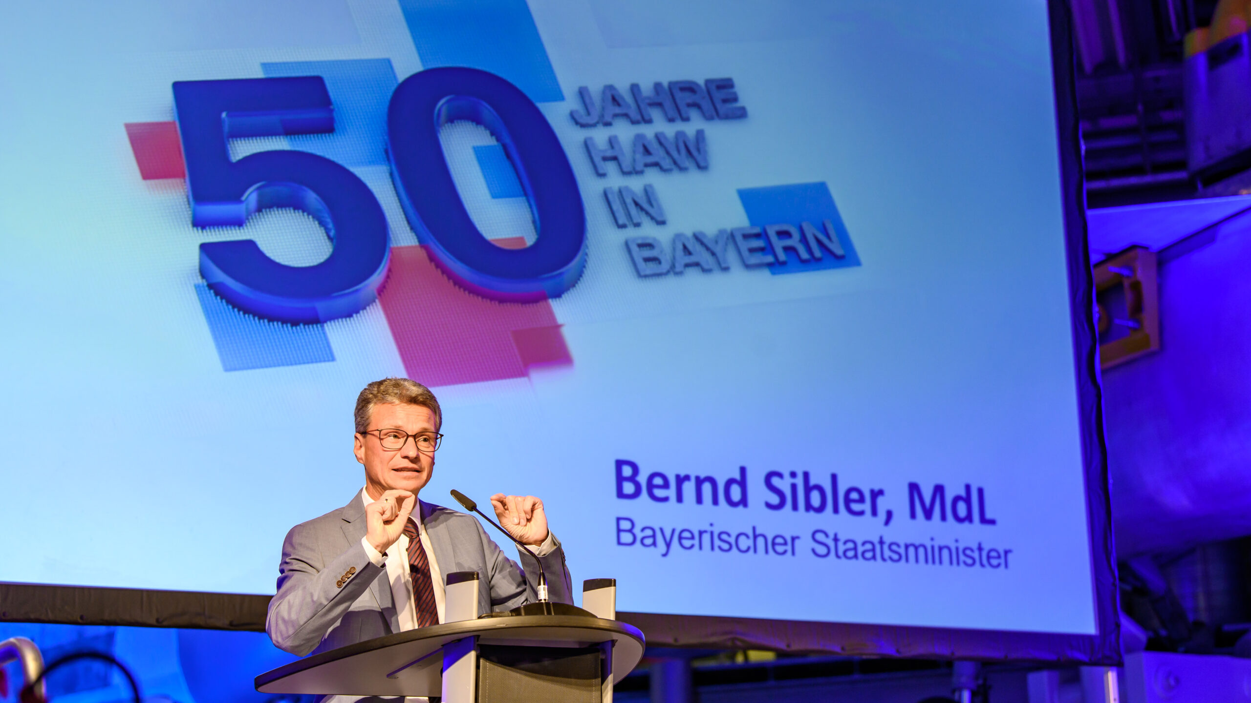 Eine Person steht an einem Podium und hält eine Rede. Hinter ihr ist ein großer Bildschirm zu sehen, auf dem „50 Jahre HAW in Bayern“ und der Text „Bernd Sibler, MdL, Bayerischer Staatsminister“ zu sehen sind. Es scheint sich um eine offizielle Veranstaltung an der Hochschule Coburg zu handeln.