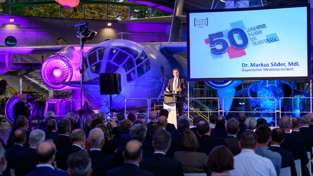 Ein Redner steht an einem Podium vor einer Menschenmenge in einem Museum, im Hintergrund ist ein historisches Flugzeug zu sehen. Auf einer großen Leinwand ist „50 Jahre in Bayern“ neben „Hochschule Coburg“ und dem Namen Dr. Markus Söder, MdL, Bayerischer Ministerpräsident zu sehen.