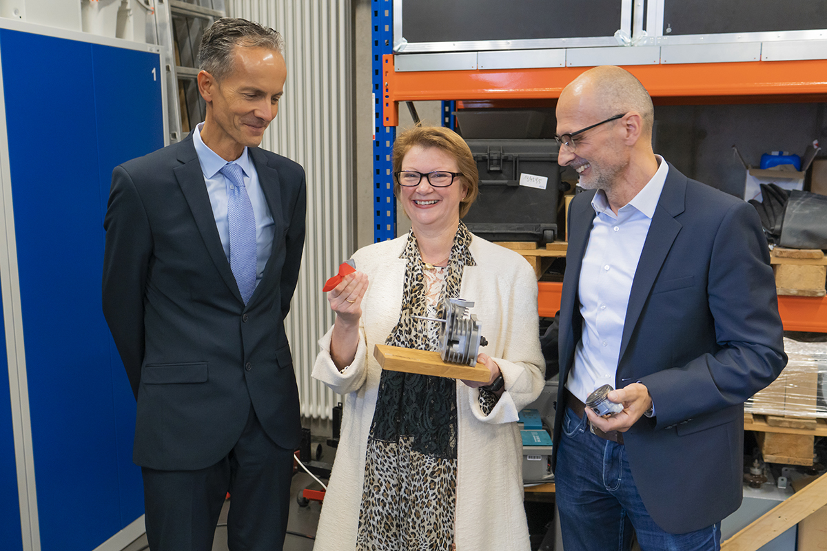 Drei Menschen stehen in einer Werkstatt der Hochschule Coburg zusammen. Die Frau in der Mitte hält lächelnd ein kleines Metallgerät und einen roten Gegenstand in der Hand. Die beiden Männer zu beiden Seiten, in Business-Kleidung gekleidet, schauen interessiert zu. Im Hintergrund sind Regale und Geräte zu sehen.