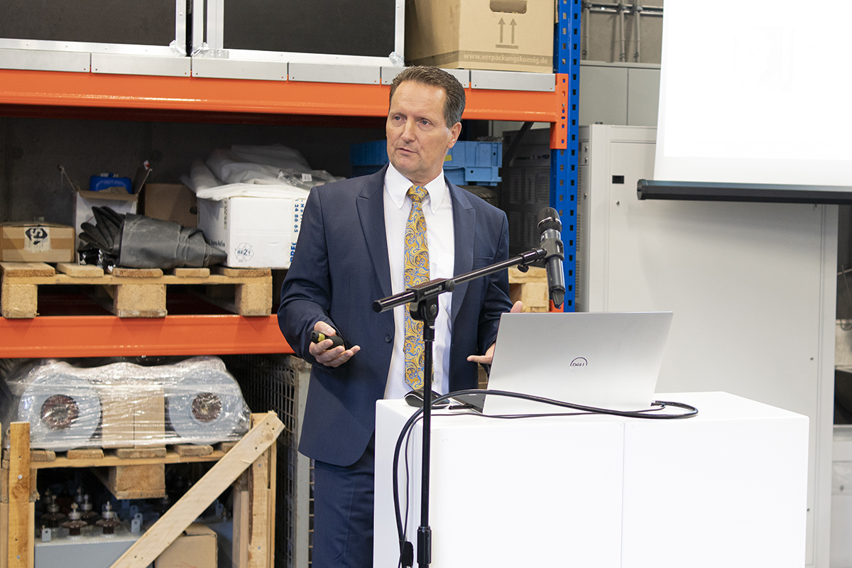 Ein Mann im Anzug hält einen Vortrag in einem Industrieumfeld und vertritt die Hochschule Coburg. Er steht mit Laptop und Mikrofon hinter einem weißen Rednerpult, im Hintergrund sind Regale mit verschiedenen Gegenständen zu sehen.