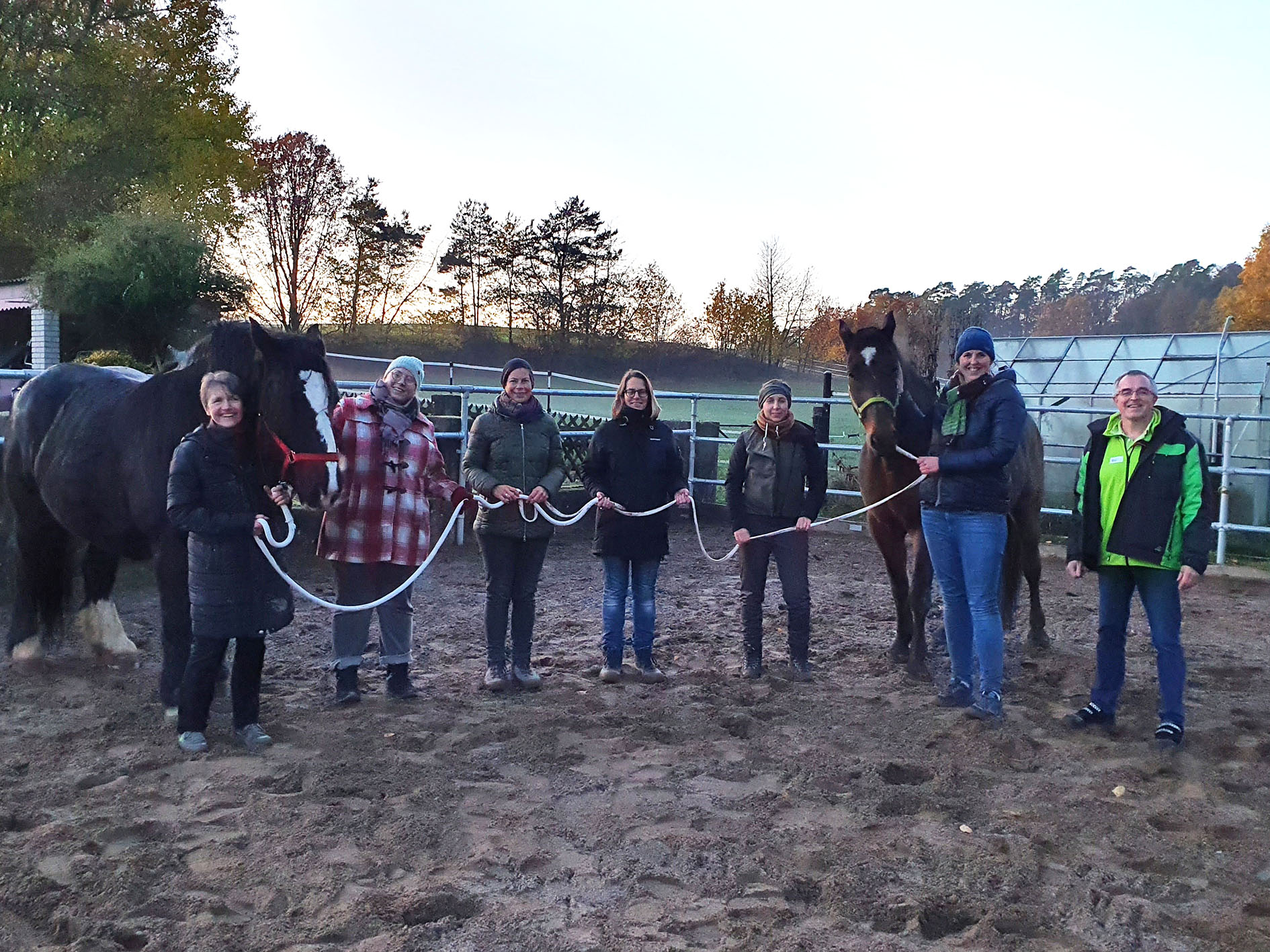 Eine Gruppe von sieben Personen, möglicherweise Studenten der Hochschule Coburg, steht im Freien auf einem Sandplatz. Jeder hält sich an Seilen fest, die an zwei großen Pferden befestigt sind. Im Hintergrund sind Bäume und ein teilweise bewölkter Himmel zu sehen, was an eine Farm- oder Ranch-Kulisse erinnert. Die Personen sind warm gekleidet.