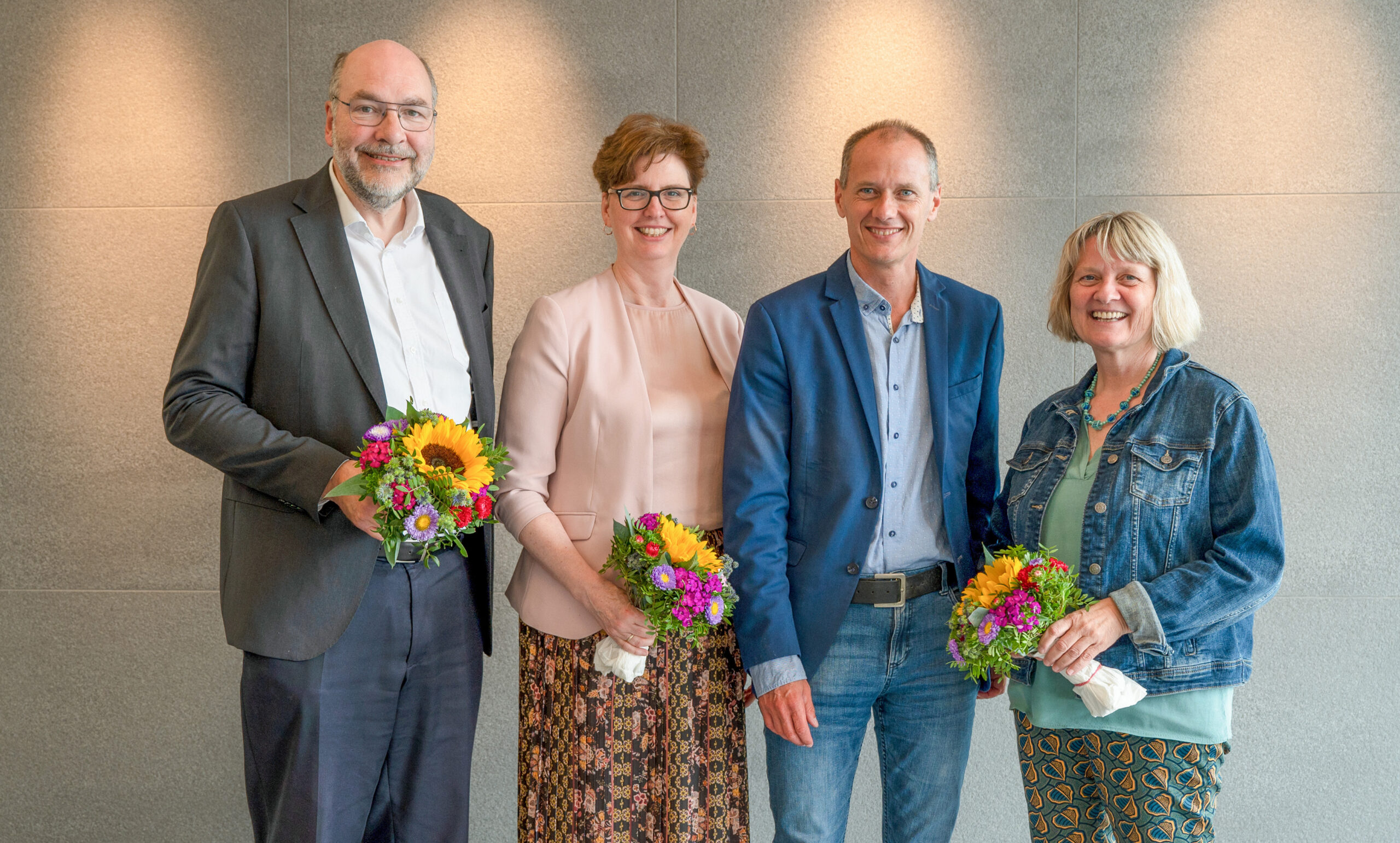 Vier Menschen stehen Seite an Seite und lächeln strahlend. Jeder hält einen bunten Blumenstrauß in der Hand. Die beiden Männer tragen Sakko, während die Frauen Bluse und Jeansjacke tragen. Sie stehen vor einem neutralen Hintergrund mit sanfter Beleuchtung – eine perfekte Momentaufnahme der Kameradschaft an der Hochschule Coburg.