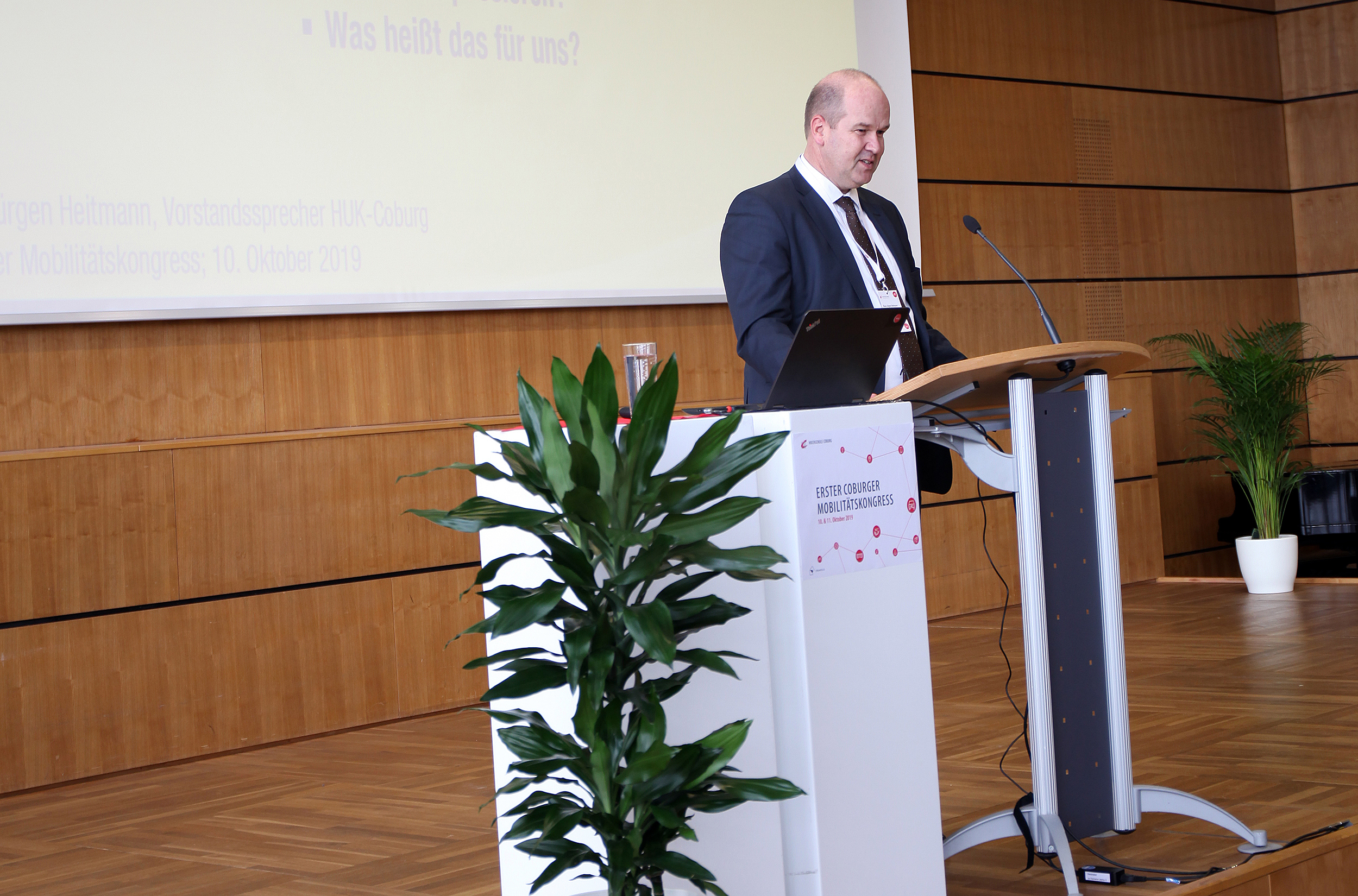 Eine Person im Anzug steht an einem Podium und hält einen Vortrag auf einer Konferenz der Hochschule Coburg. Der mit Holzvertäfelung und einer grünen Pflanze geschmückte Raum bietet ein professionelles Ambiente. In der Nähe zeigt ein Bildschirm Text auf Deutsch.