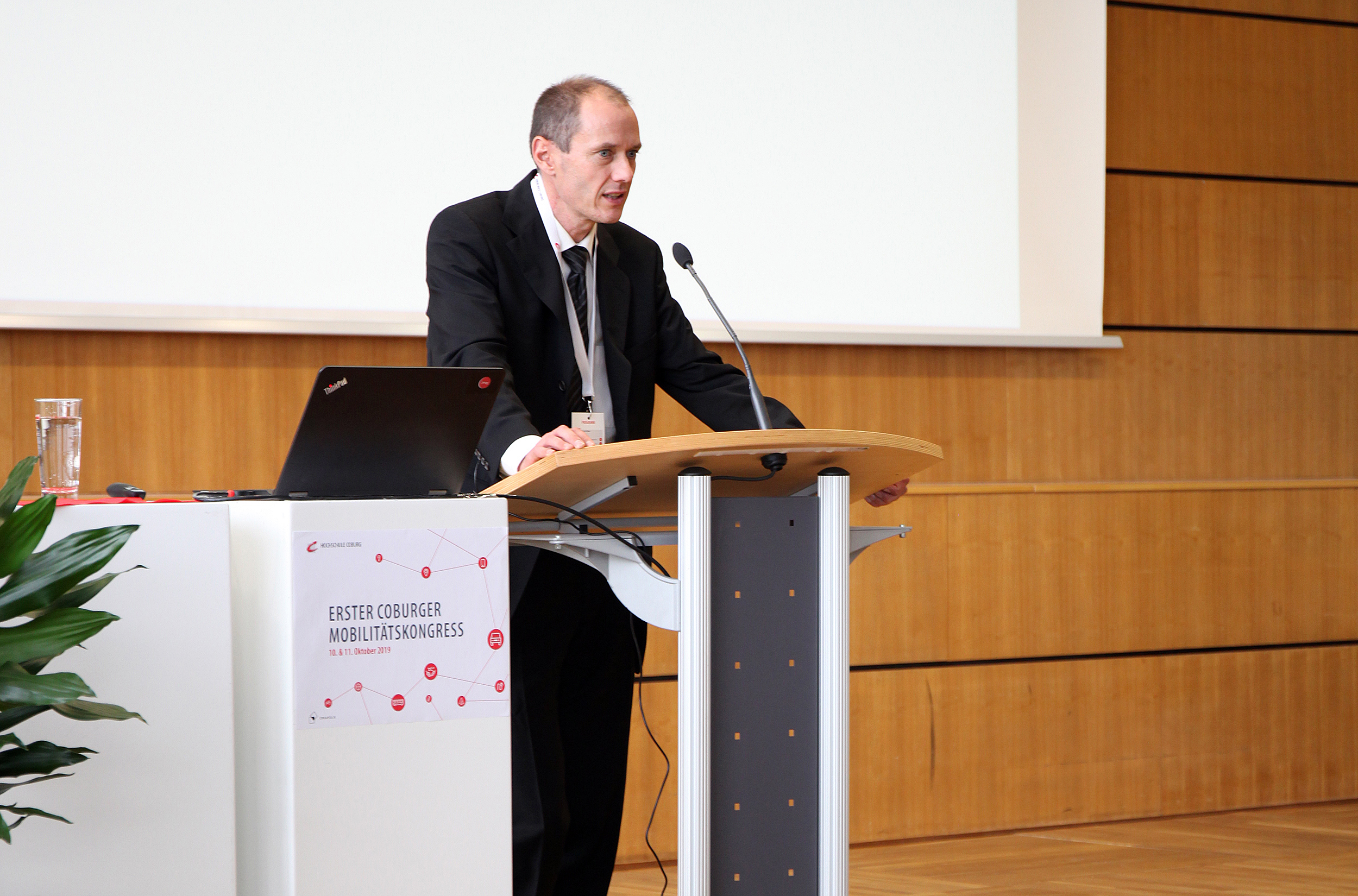 Ein Mann im Anzug steht an einem Podium in einem Konferenzsaal und spricht in ein Mikrofon. Auf dem Podium stehen ein Laptop und ein Glas Wasser. Auf einem Plakat steht „Erster Coburger Mobilitätskongress“, stolz präsentiert von der Hochschule Coburg.