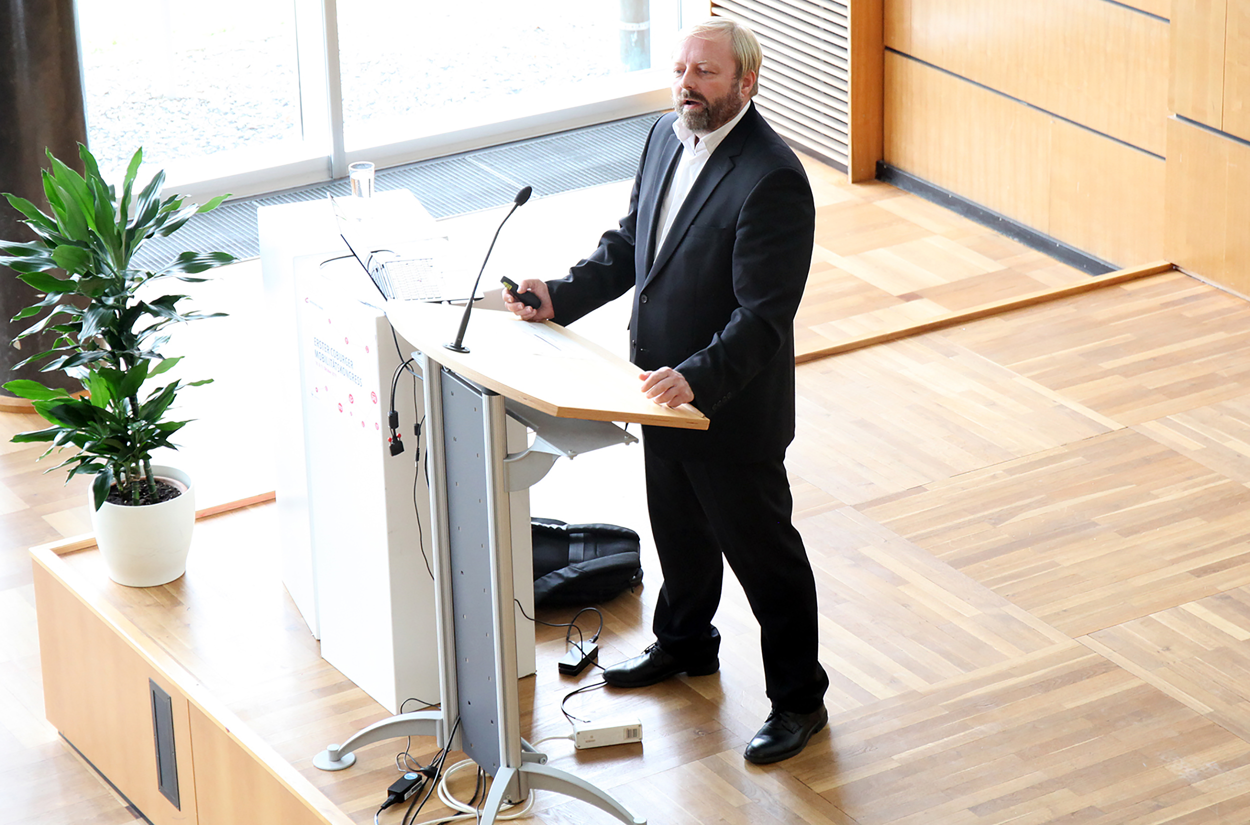 Ein Mann im Anzug steht an einem Podium und hält einen Vortrag an der Hochschule Coburg in einem hellen Raum mit Holzboden. Er hält ein Mikrofon in der Hand und wendet sich an das Publikum. Links von ihm ist eine Pflanze in einem weißen Topf zu sehen.