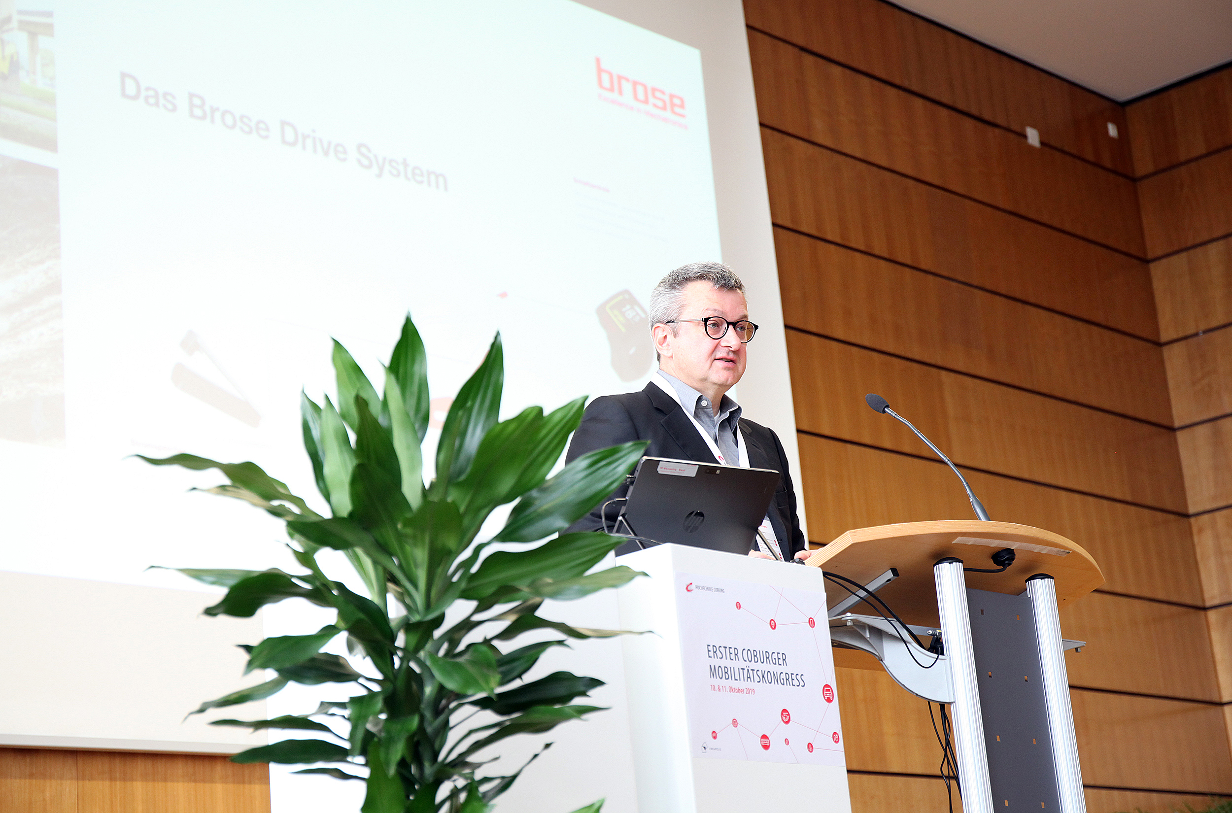 Ein Mann steht hinter einem Podium und hält einen Vortrag an der Hochschule Coburg. Im Hintergrund ist auf einer großen Leinwand ein Teil des Textes „Das Brose Antriebssystem“ zu sehen. Eine Topfpflanze schmückt die linke Seite des Podiums und ergänzt das professionelle Ambiente des Konferenzraums.