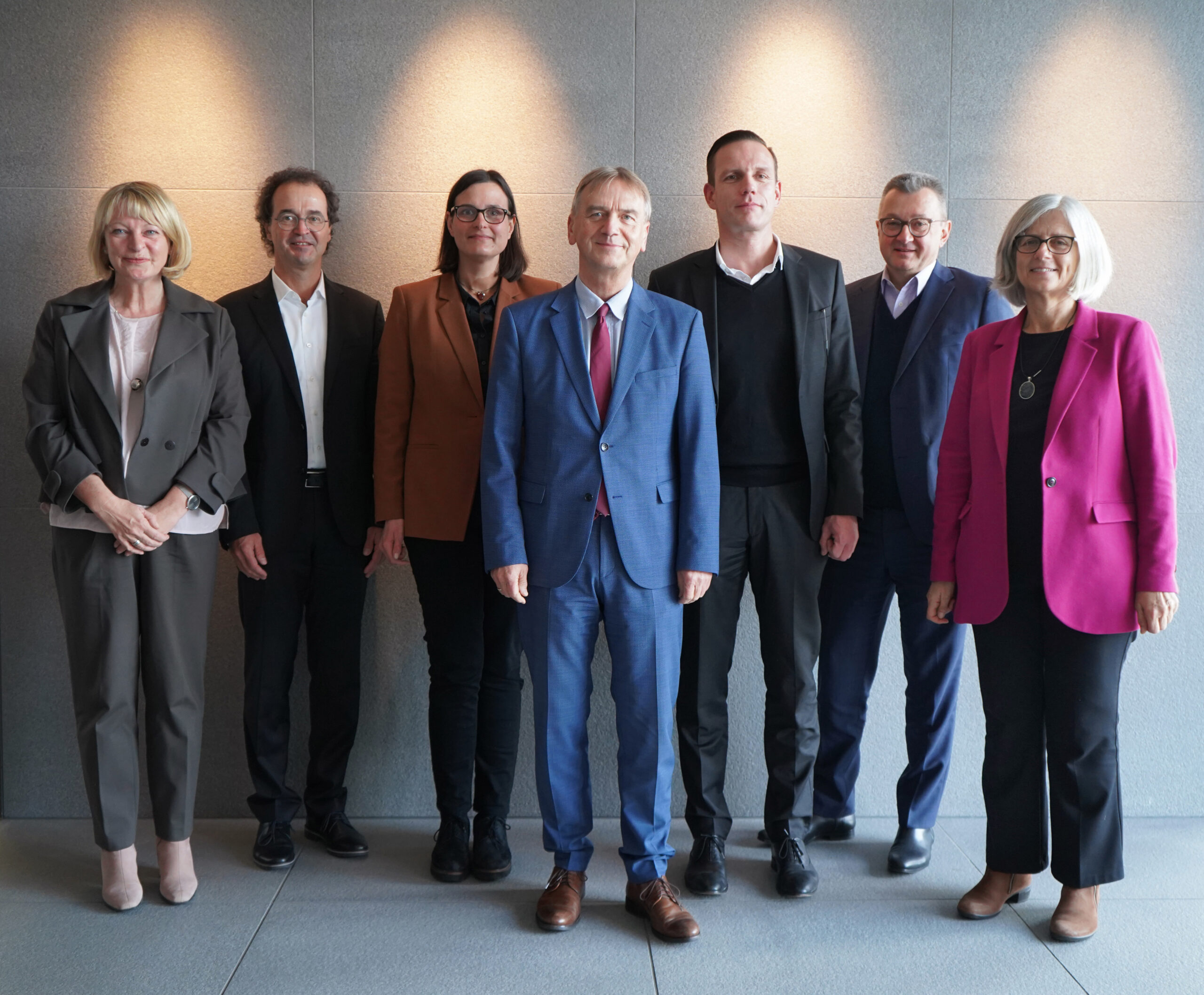 Eine Gruppe von sieben Personen steht in einem Innenbereich der Hochschule Coburg vor einer grauen Wand. Fünf von ihnen sind in professioneller Kleidung gekleidet, fünf tragen Sakko über Hemden, während zwei Frauen leuchtende Blazer tragen – einer rosa, einer orange – und selbstbewusst in die Kamera blicken.