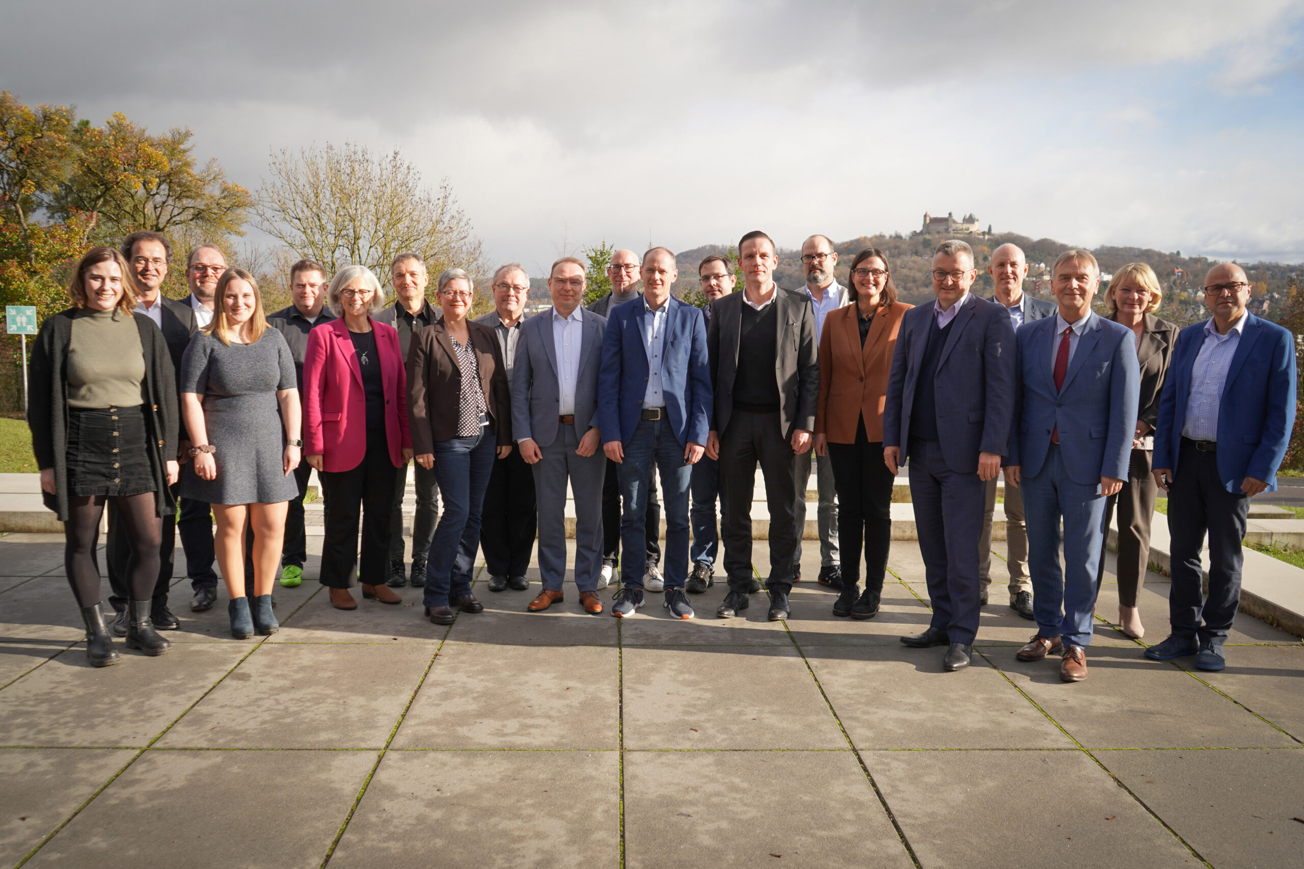 Eine Gruppe von 22 Personen, sowohl Männer als auch Frauen, steht draußen auf einer gepflasterten Fläche in der Nähe der Hochschule Coburg. Sie sind in Business- und Freizeitkleidung gekleidet. Hinter ihnen ist eine Landschaft mit Bäumen und einem bewölkten Himmel zu sehen.