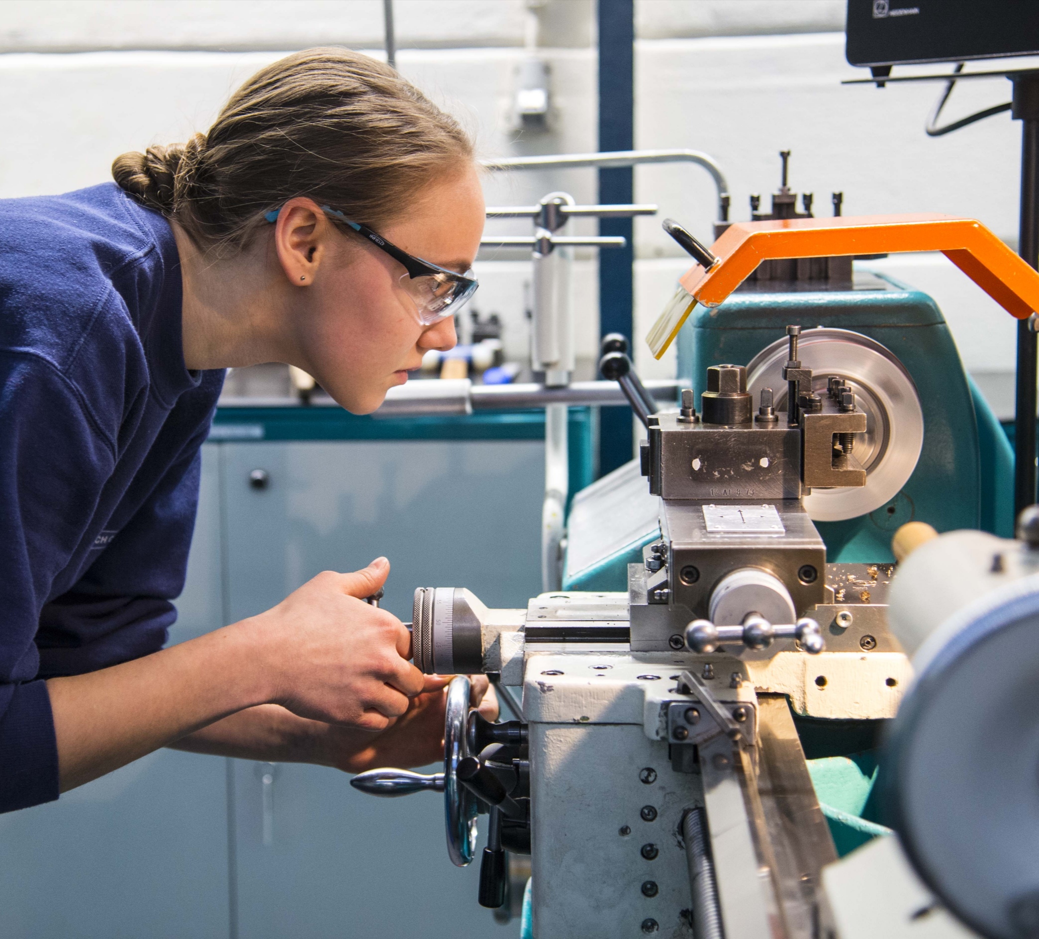 In der Industriewerkstatt der Hochschule Coburg bedient eine Person mit Schutzbrille eine Drehmaschine. Die Person ist konzentriert auf die Einstellung der Maschine, umgeben von verschiedenen mechanischen Komponenten. Die Umgebung ist ausgesprochen technisch und auf Präzision ausgerichtet.